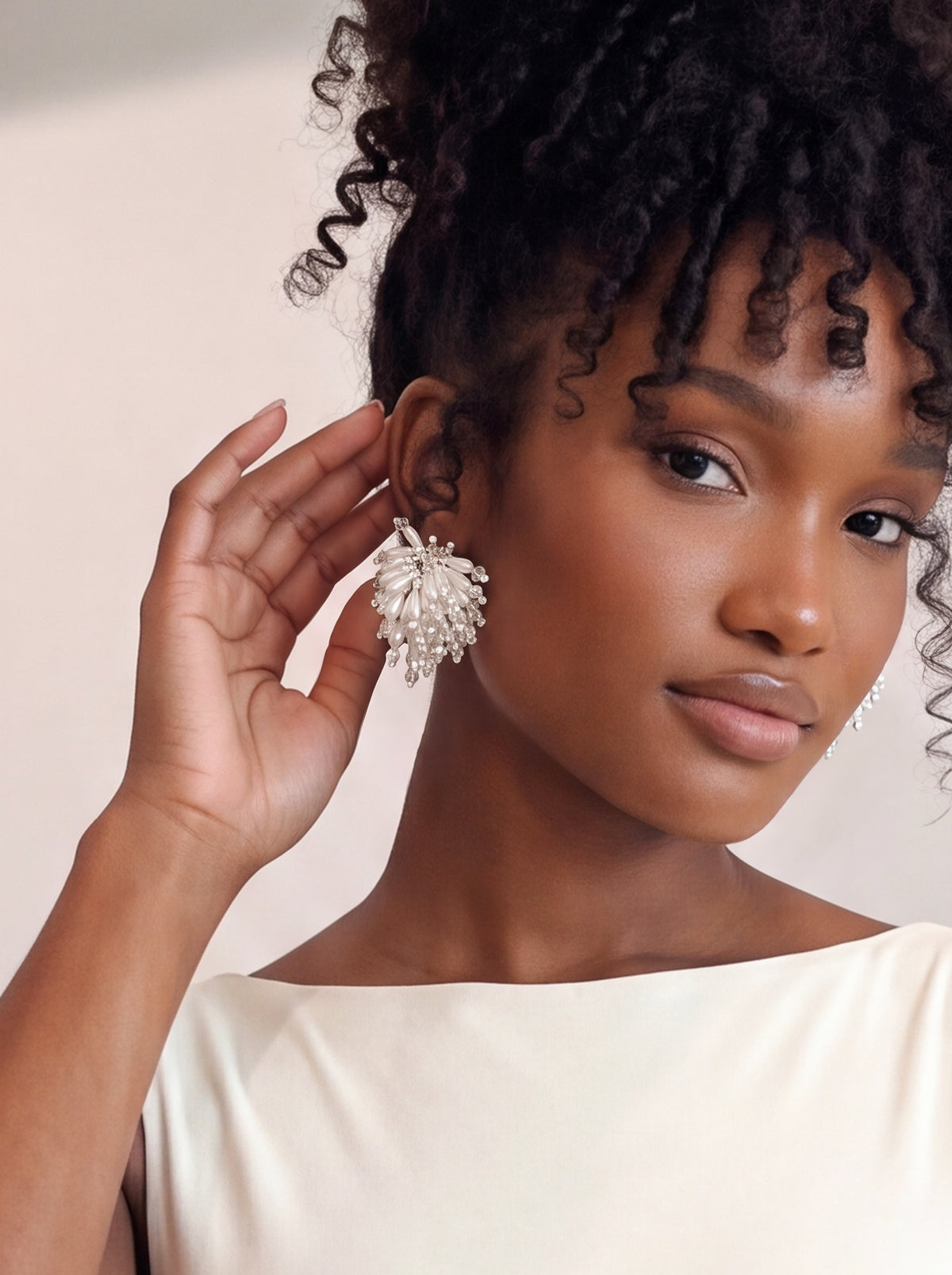 Close-up of a model wearing oversized ivory floral statement earrings featuring layered pearl-like petals, shimmering crystal beads, and cascading pearl-inspired embellishments. Styled with an ivory sleeveless top and voluminous curly updo, gently touching her ear to highlight the earrings’ intricate 3D design and elegant bridal-inspired detailing.