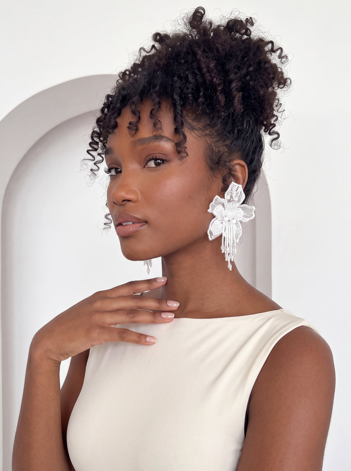 Close-up of a model wearing oversized white floral lace statement earrings featuring intricate embroidered petals, delicate pearl-like accents, and dangling beaded tassels. Styled with an ivory sleeveless bodysuit and voluminous curly updo against a minimalist white background.