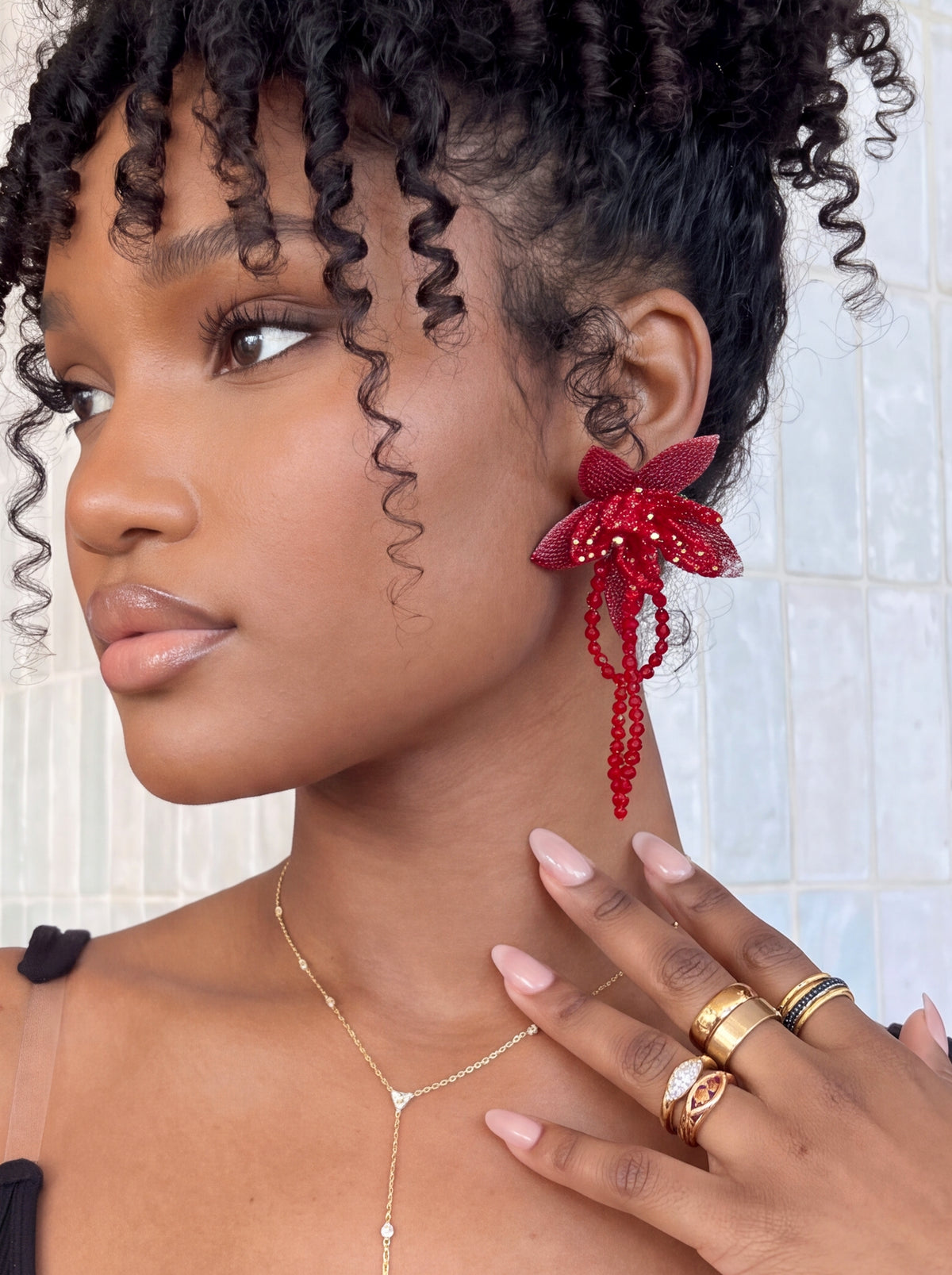 A close-up lifestyle shot of a model wearing a vibrant red beaded flower-shaped dangle earring, gold rings, and a delicate gold necklace against a light tiled wall.