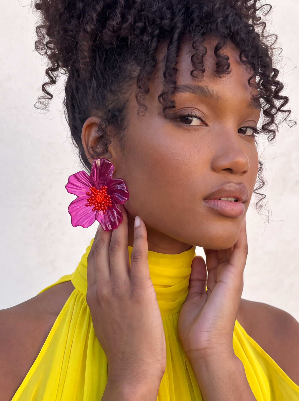 A close-up portrait of a woman with curly dark hair wearing a bright yellow high-neck top and a large magenta flower-shaped earring with a red center, posing against a light neutral background.
