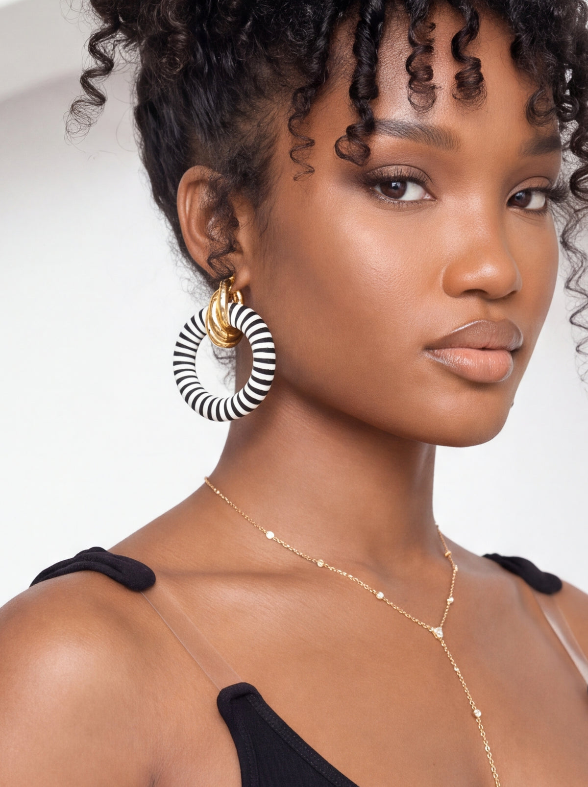 A close-up of a model wearing a pair of large black and white striped hoop earrings with gold accents, along with a delicate gold necklace.