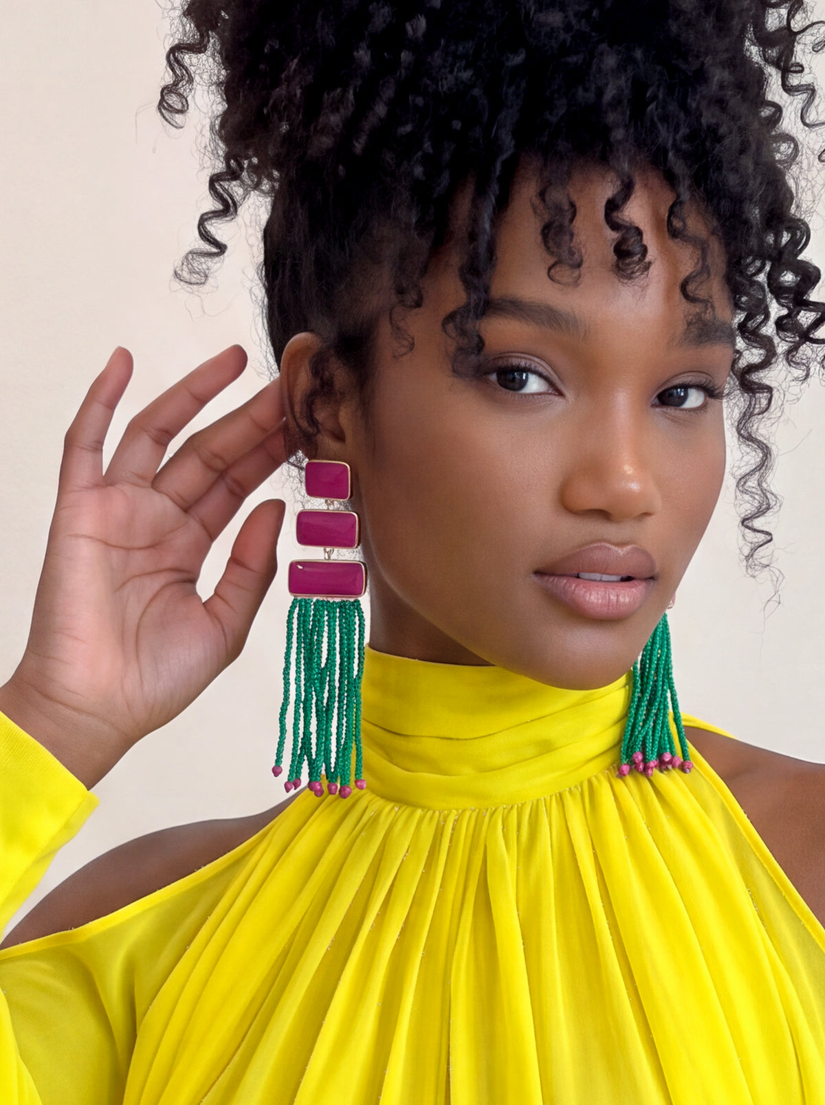 A close-up of a model wearing a pair of large statement earrings. The earrings feature three stacked magenta-pink rectangular beads on a silver-tone wire frame, with long green beaded tassels hanging below.