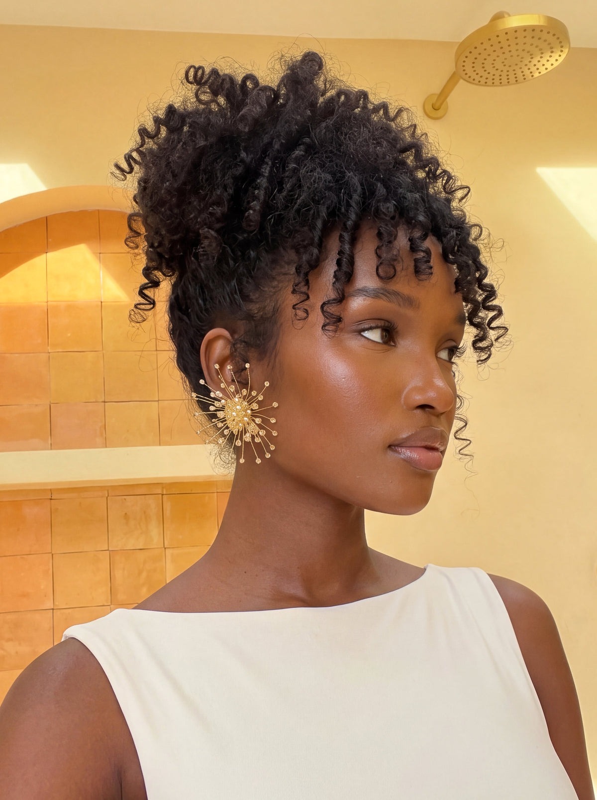  Close-up of a model wearing oversized gold starburst statement earrings featuring delicate radiating wires and shimmering bead accents. Styled with a white sleeveless top and voluminous curly updo, highlighting the earrings’ intricate sculptural design and elegant gold finish.