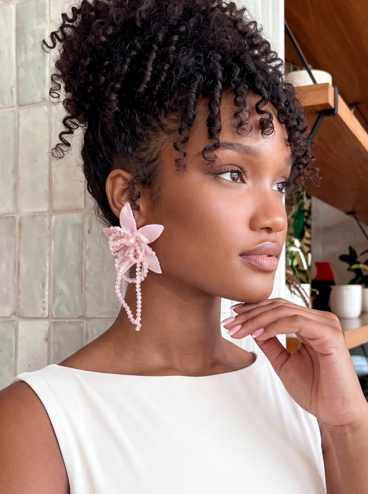 Close-up of a model wearing oversized blush pink floral statement earrings featuring delicate embroidered mesh petals, intricate 3D beadwork, and cascading pink beaded tassels. Styled with an ivory sleeveless top and voluminous curly updo, highlighting the earrings’ romantic floral design and soft pink detailing.
