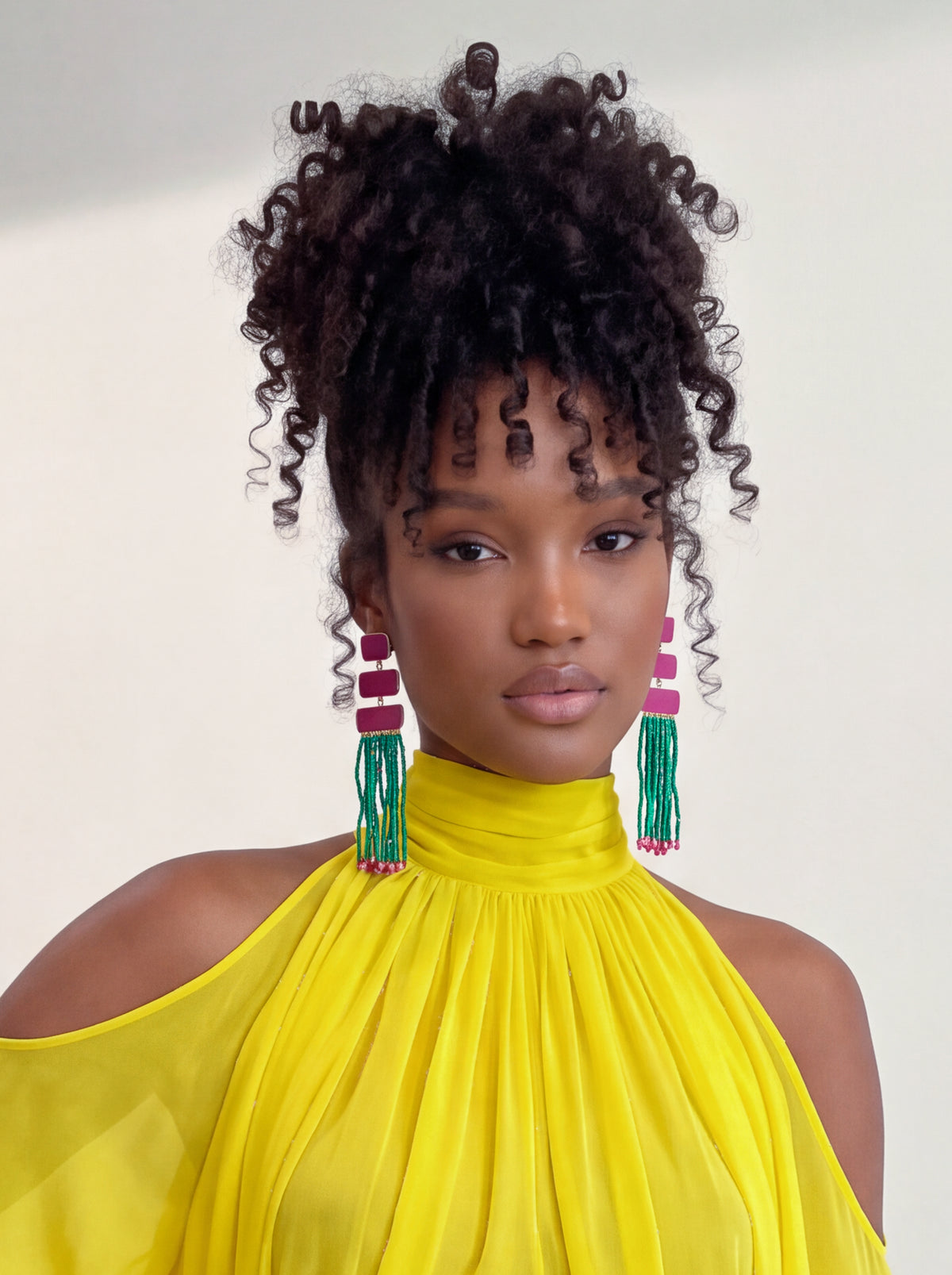 A woman with voluminous dark curly hair styled in a high puff bun, wearing a bright yellow high-neck cold-shoulder top and colorful dangling earrings, set against a plain off-white background.