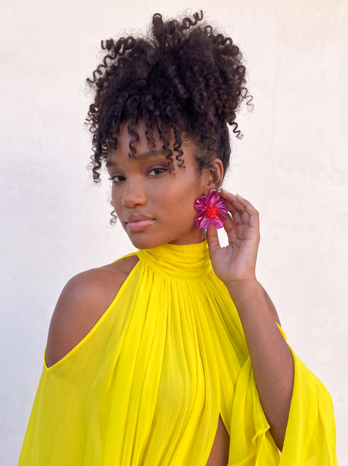 A woman with curly black hair wearing a bright yellow high-neck cold-shoulder top and a large pink flower earring, posing against a light beige wall.