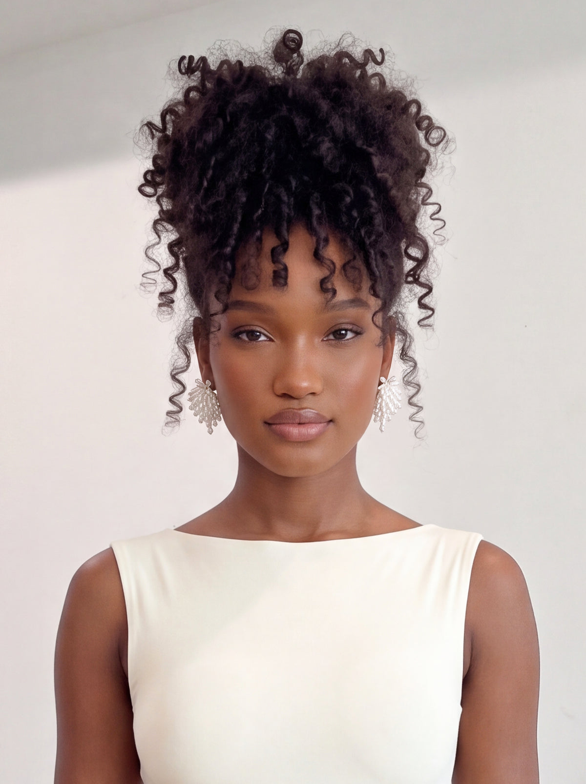 Front-facing model wearing oversized ivory floral statement earrings featuring layered pearl-inspired petals, shimmering crystal bead accents, and delicate cascading beadwork. Styled with a sleek ivory sleeveless top and voluminous curly updo against a minimalist white background, showcasing the earrings’ elegant, dimensional design.