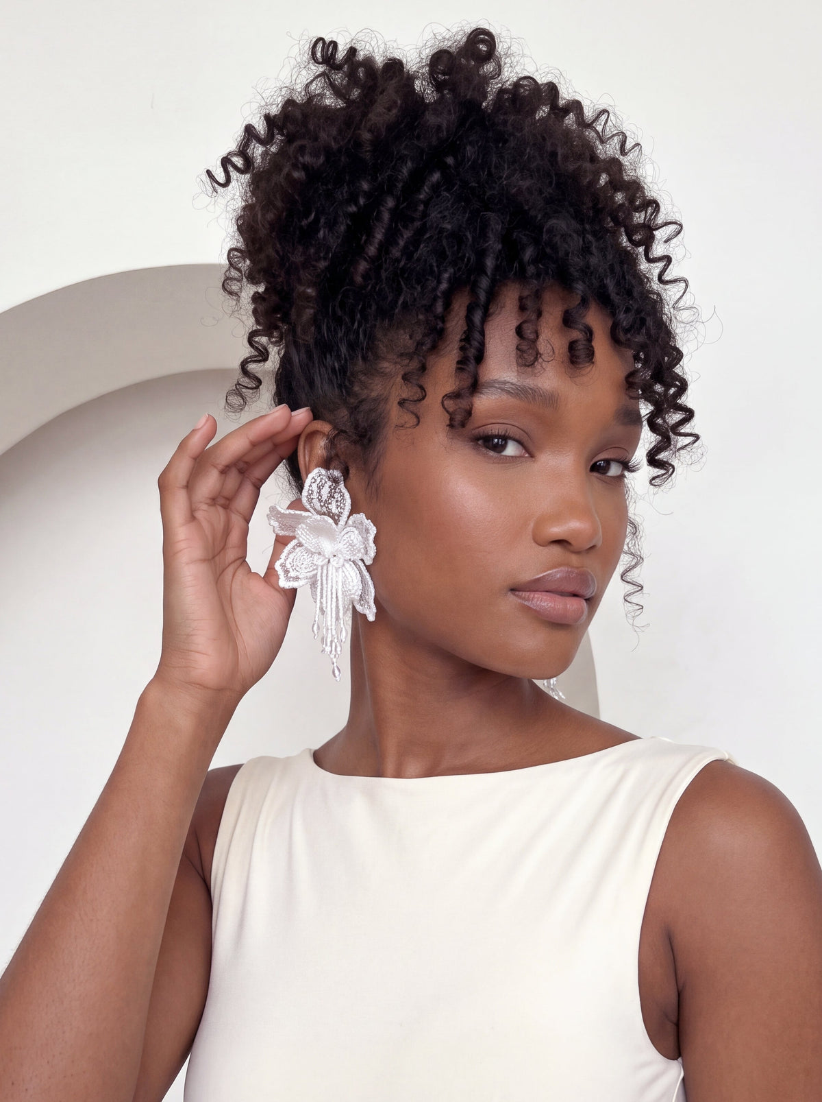 Model wearing elegant white 3D floral lace statement earrings with layered embroidered petals and cascading beaded fringe. Styled with a fitted ivory sleeveless bodysuit and curly updo, gently touching the earring to highlight its intricate detailing and romantic, feminine design.