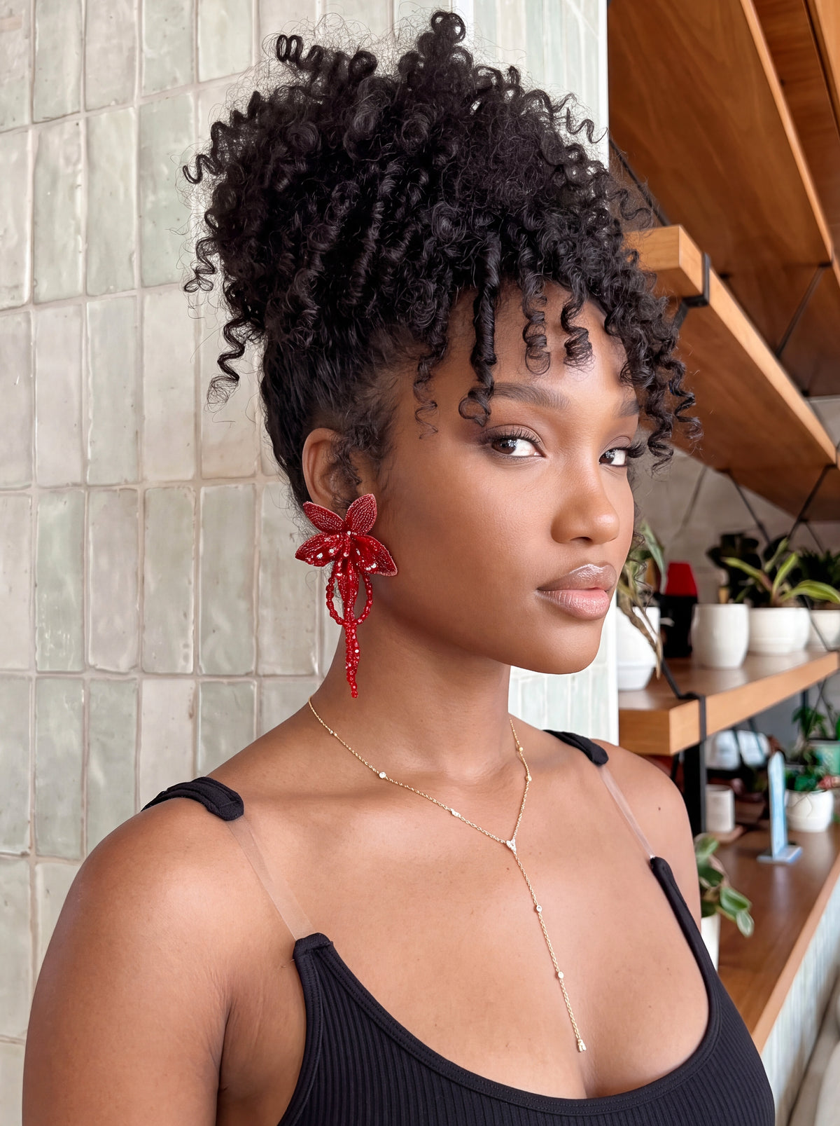 A woman with voluminous, tightly curled black hair is shown in a close-up portrait. She is wearing a black ribbed tank top, a delicate gold necklace, and a prominent red beaded flower-shaped earring.
