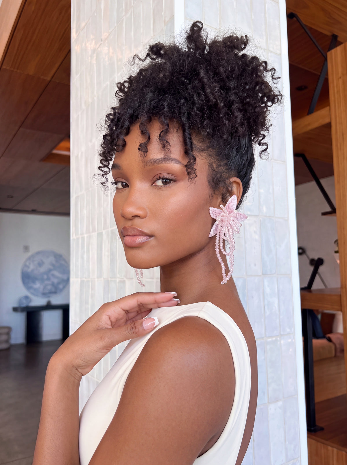 Model wearing oversized blush pink floral statement earrings featuring delicate embroidered mesh petals, intricate 3D beadwork, and long cascading pink beaded tassels. Styled with an ivory sleeveless bodysuit and voluminous curly updo, posing sideways to highlight the earrings’ romantic floral silhouette and elegant dangling details.