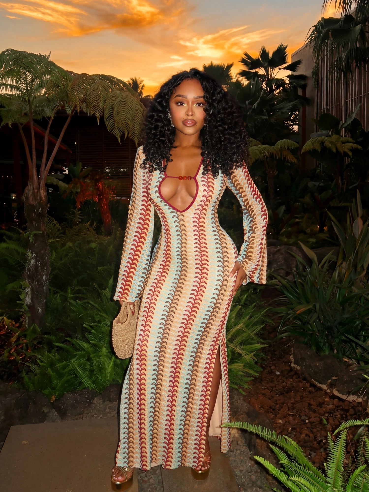 Woman wearing a fitted crochet maxi dress with long sleeves and front cutout standing in a tropical garden at sunset with palm trees and warm sky behind her.