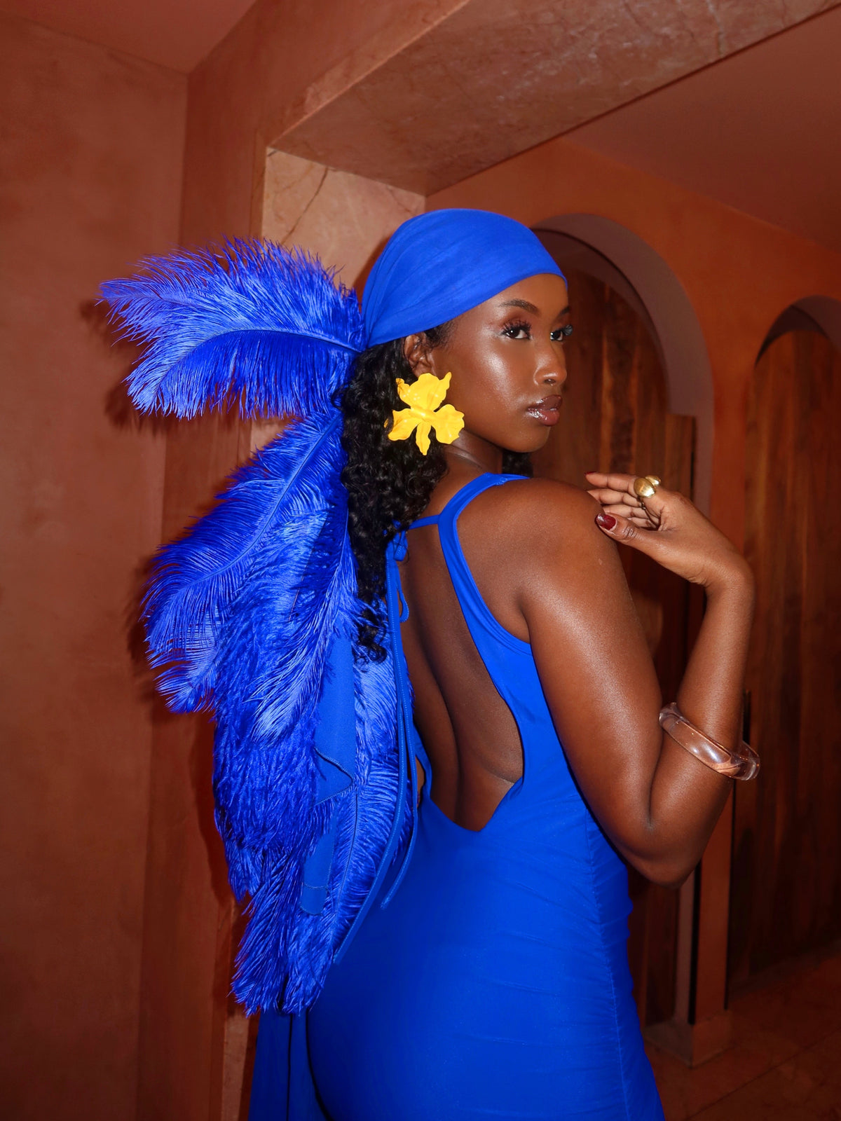 A woman wearing a bright royal blue sleeveless dress, matching blue headwrap, and large blue feathered shoulder accessories, posed indoors against warm terracotta walls.