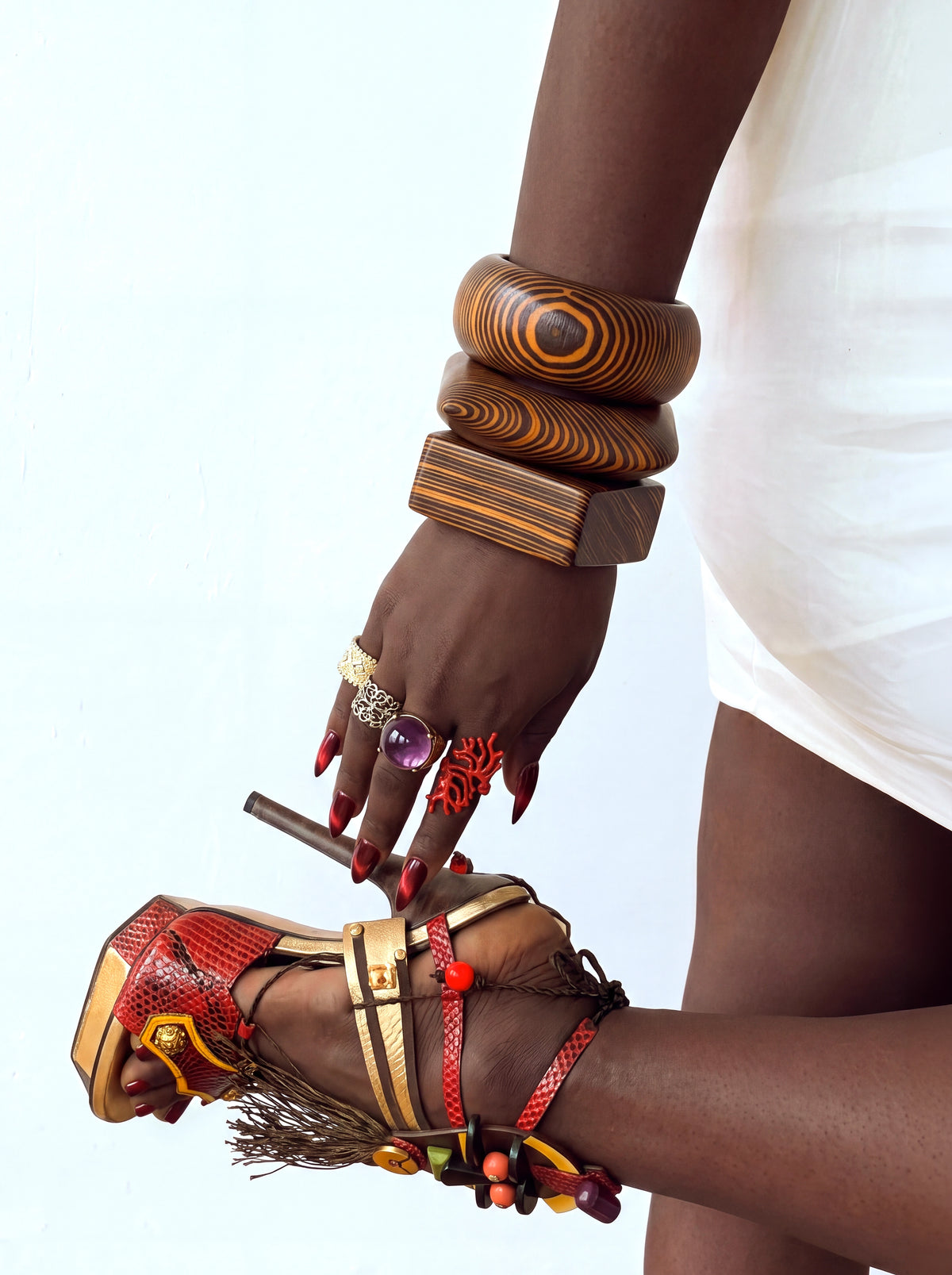 Close-up fashion shot featuring stacked oversized wood-grain bangles, colorful statement rings, metallic burgundy nails, and embellished strappy heels with beaded and tassel details, styled with a white mini dress.