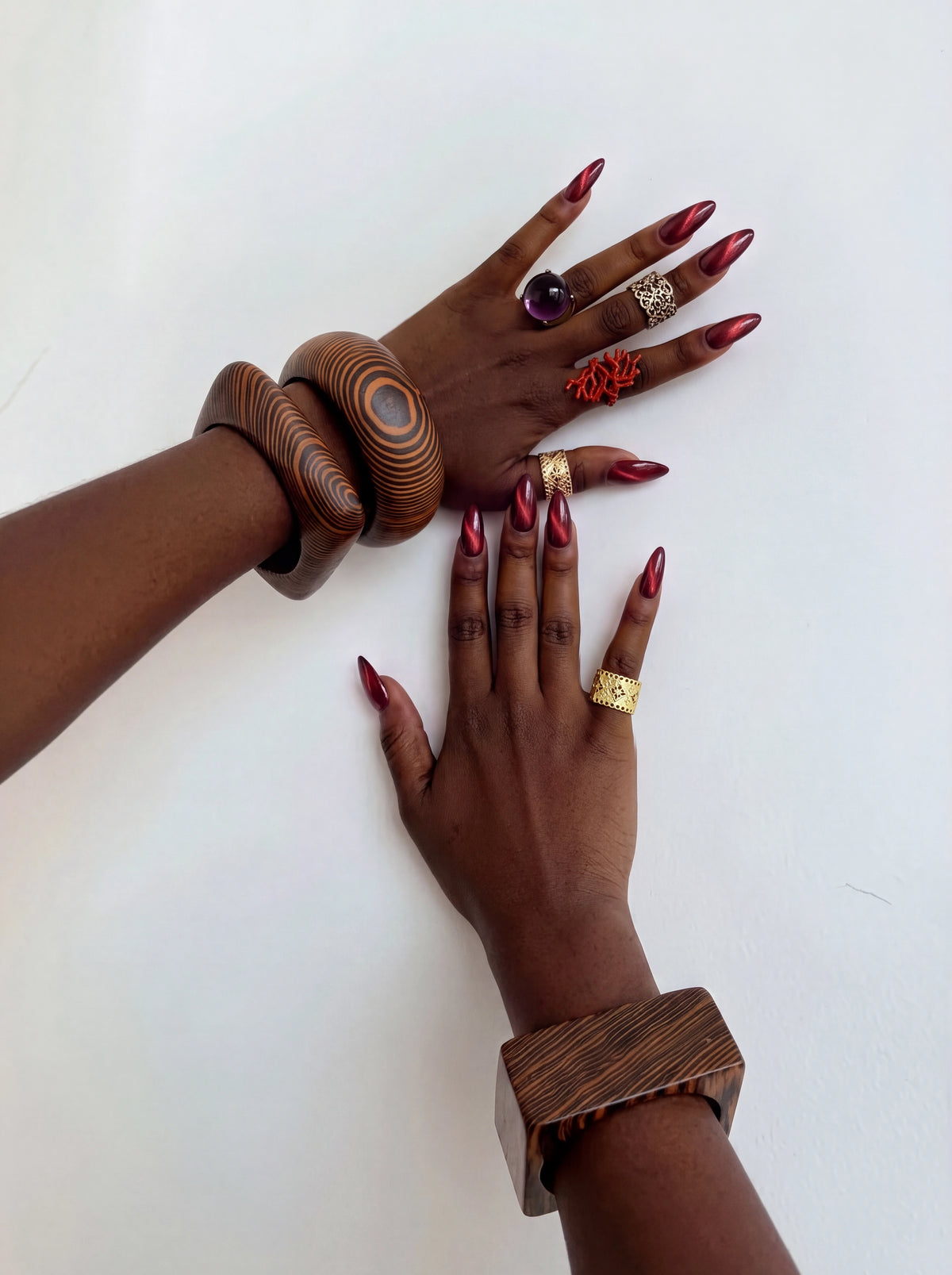 Two hands styled with bold statement jewelry, featuring oversized wood-grain bangles, gold and colorful rings, and deep burgundy metallic almond-shaped nails against a clean white background.