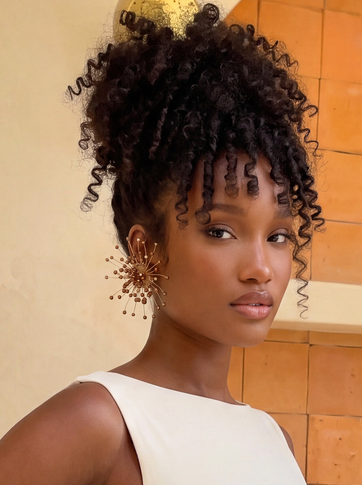 Close-up of a model wearing oversized bronze and gold starburst statement earrings featuring delicate radiating wires and shimmering bronze bead accents. Styled with a sleek ivory sleeveless top and voluminous curly updo, showcasing the earrings’ intricate 3D sculptural design and warm metallic finish.