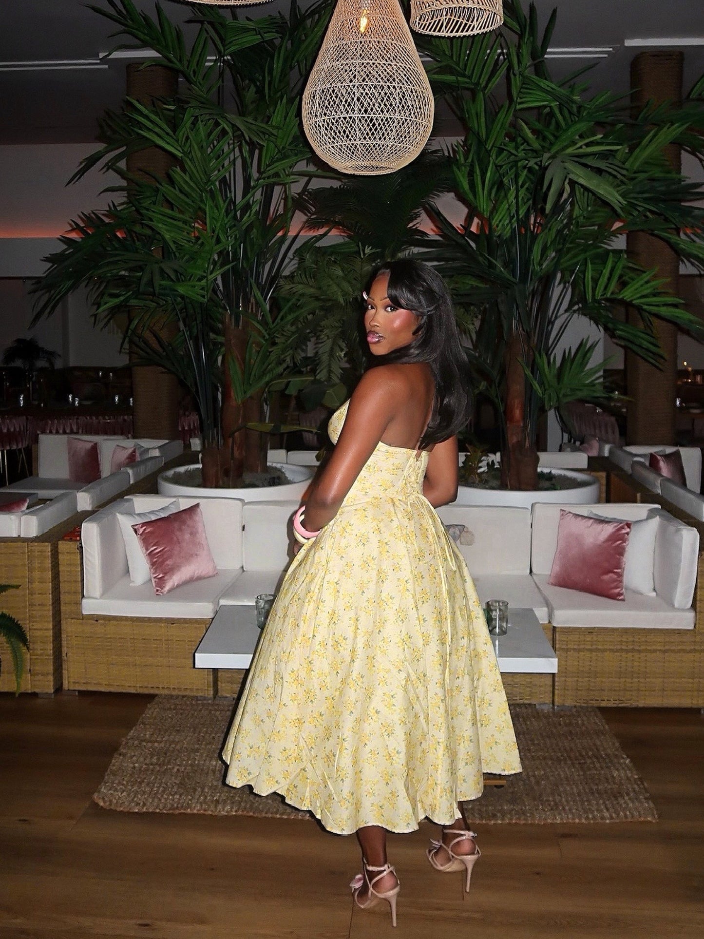 Back view of a yellow floral halter midi dress featuring a fitted waist, smocked back, and full flowing skirt. Styled with floral heels and pastel bangles, the model poses in a tropical lounge with palm trees and woven pendant lighting.