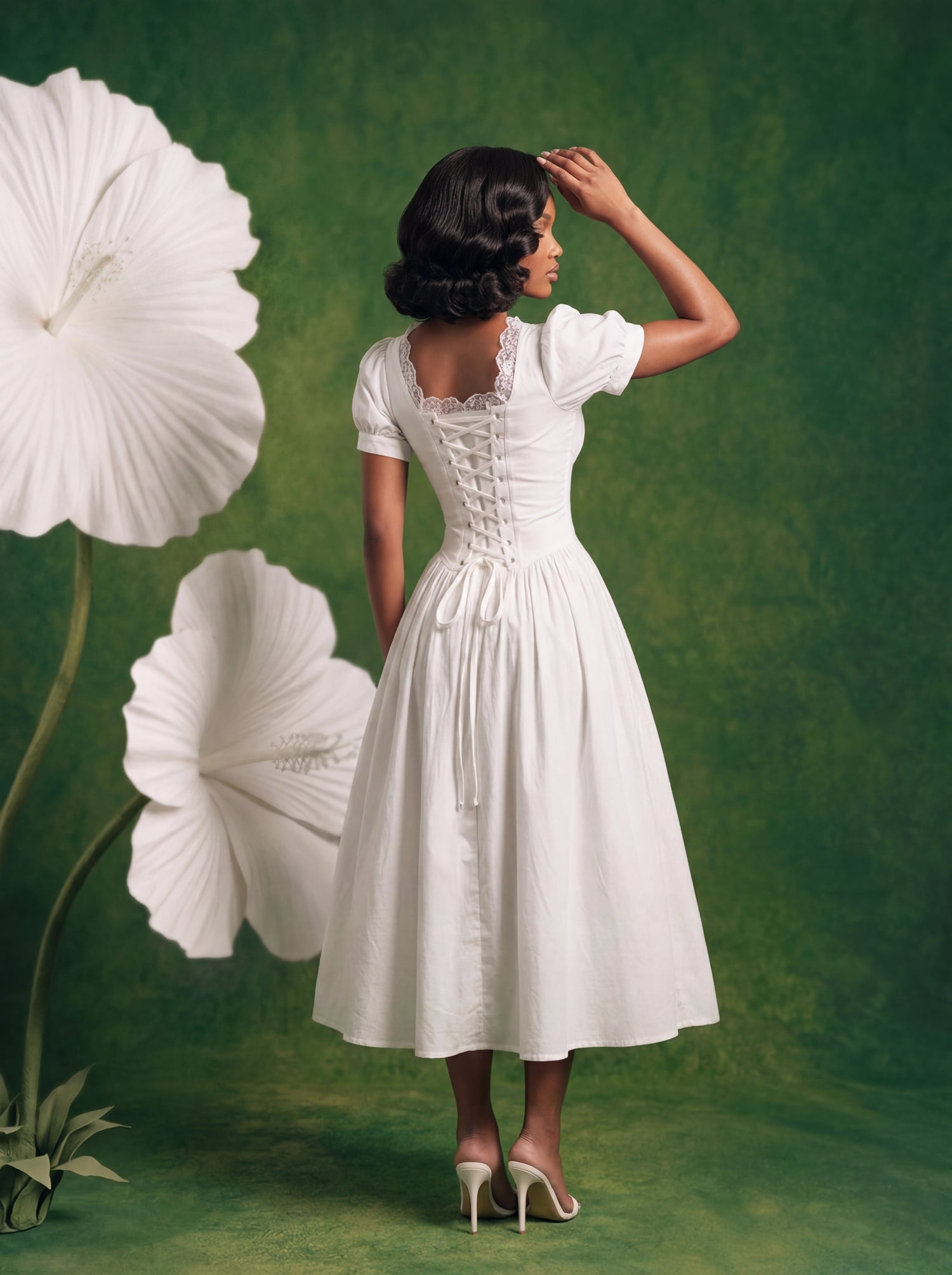 Back view of a white midi dress featuring an adjustable lace-up corset back, lace trim detailing, puff sleeves, and a flowing full skirt. Styled with white heels against a textured green backdrop with oversized floral props.