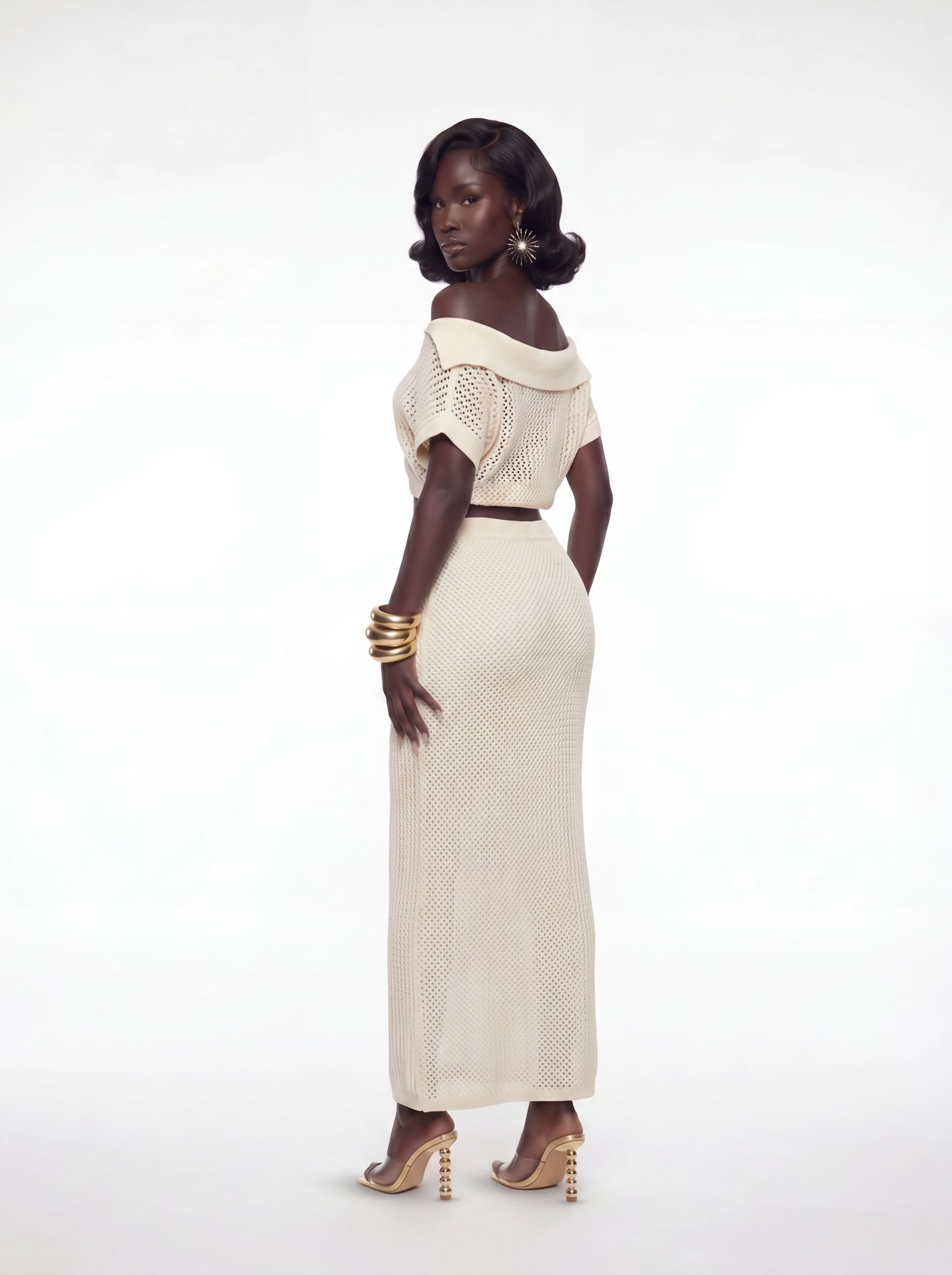 Back angled view of a cream crochet two-piece maxi skirt set featuring an off-shoulder knit crop top and fitted high-waisted crochet skirt styled with gold accessories and heels.