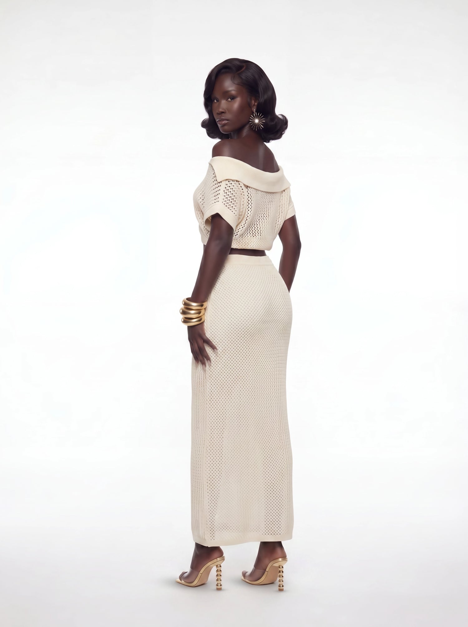 Back angled view of a cream crochet two-piece maxi skirt set featuring an off-shoulder knit crop top and fitted high-waisted crochet skirt styled with gold accessories and heels.