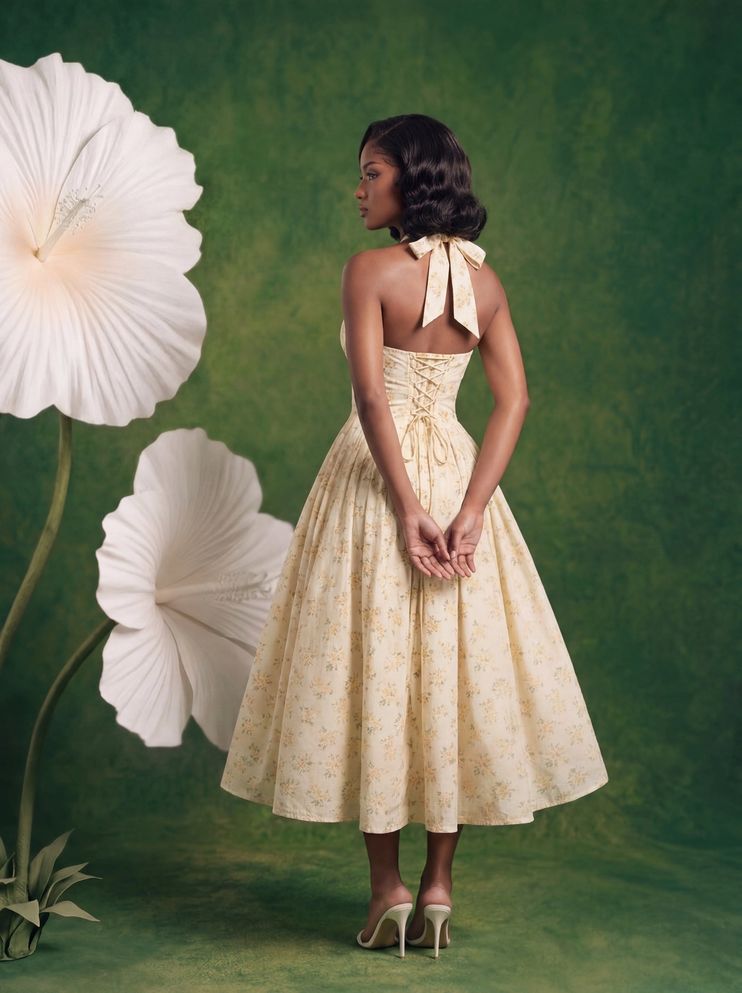 Back view of a soft yellow floral halter midi dress featuring an adjustable lace-up corset back, open-back silhouette, and full flowing skirt. Styled against a textured green backdrop with oversized white flowers for an elegant feminine aesthetic.