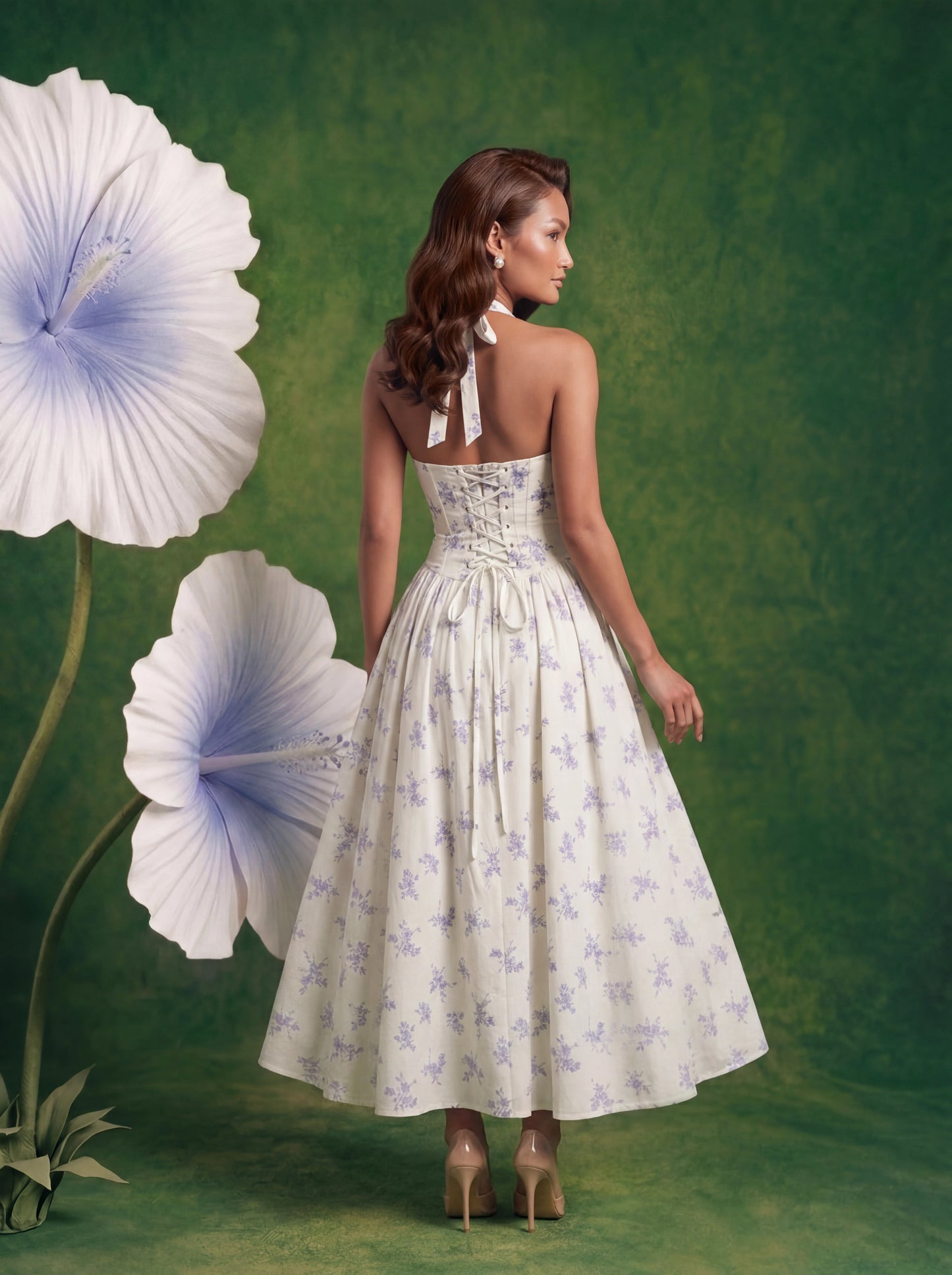 Back view of a woman with wavy warm-brunette hair standing in a surreal studio setting. She is wearing a white halter-neck midi dress featuring a delicate light blue floral print, a ruched bodice with a tie string, and a full, flared skirt. She is wearing nude open-toe heels. She is surrounded by oversized, sculptural white flowers with light blue centers and green stems, all set against a textured, earthy dark green backdrop.