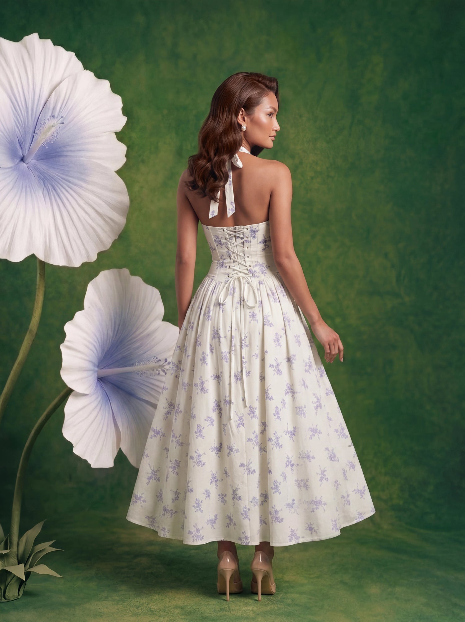 Back view of a woman with wavy warm-brunette hair standing in a surreal studio setting. She is wearing a white halter-neck midi dress featuring a delicate light blue floral print, a ruched bodice with a tie string, and a full, flared skirt. She is wearing nude open-toe heels. She is surrounded by oversized, sculptural white flowers with light blue centers and green stems, all set against a textured, earthy dark green backdrop.