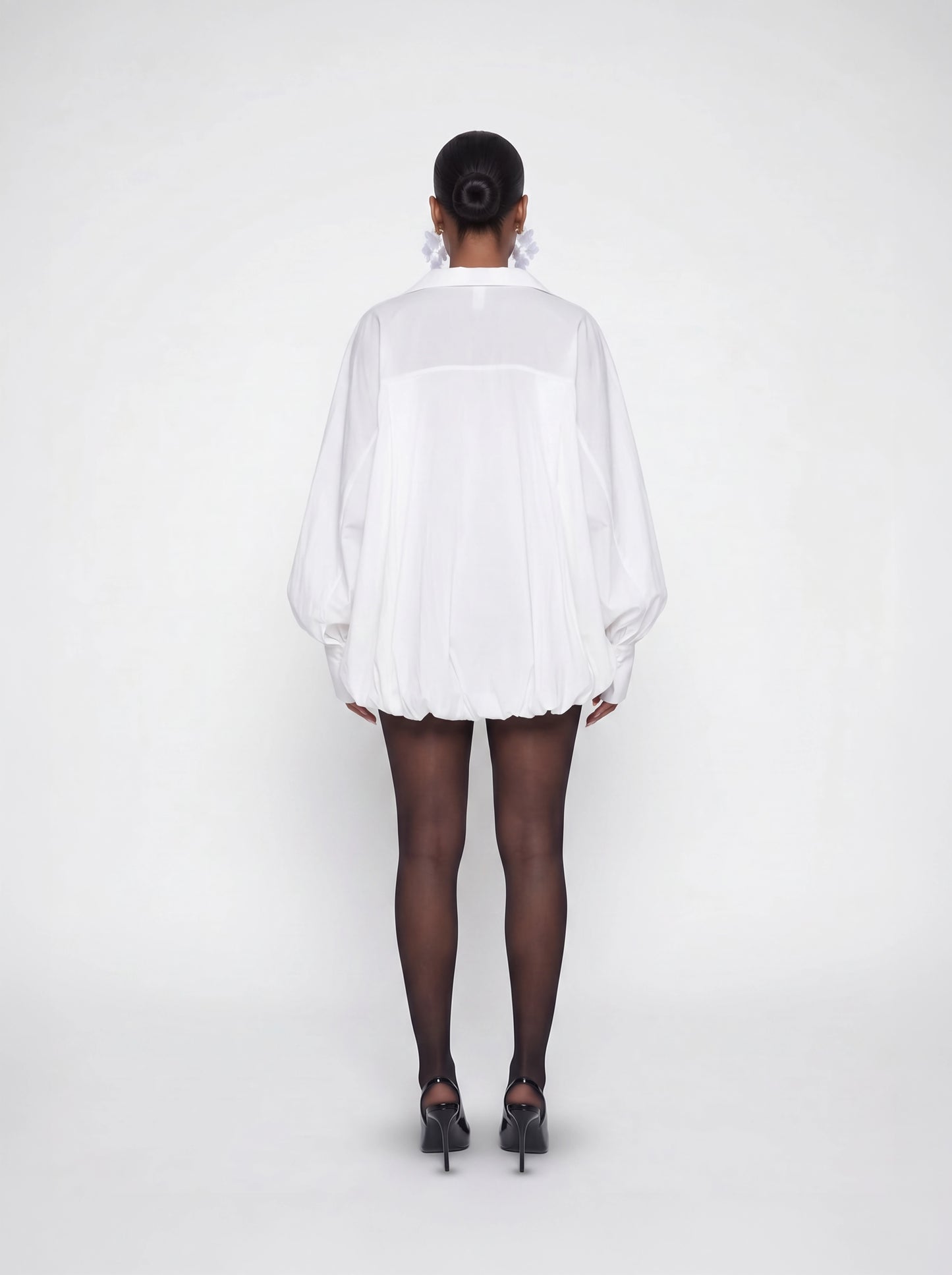 Back view of a white oversized bubble hem mini dress featuring voluminous sleeves, relaxed oversized fit, and lightweight flowy fabric styled with black heels.
