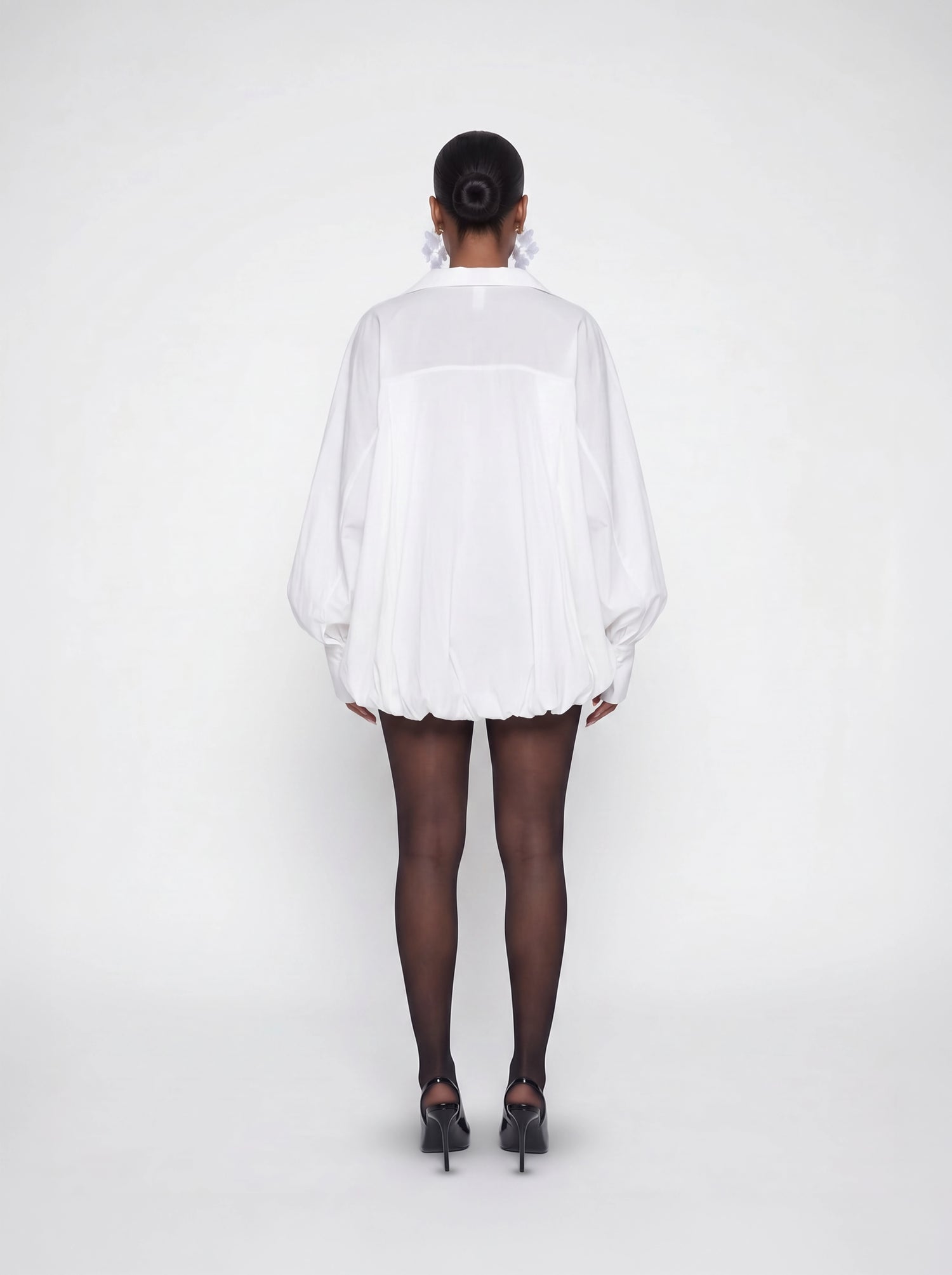 Back view of a white oversized bubble hem mini dress featuring voluminous sleeves, relaxed oversized fit, and lightweight flowy fabric styled with black heels.