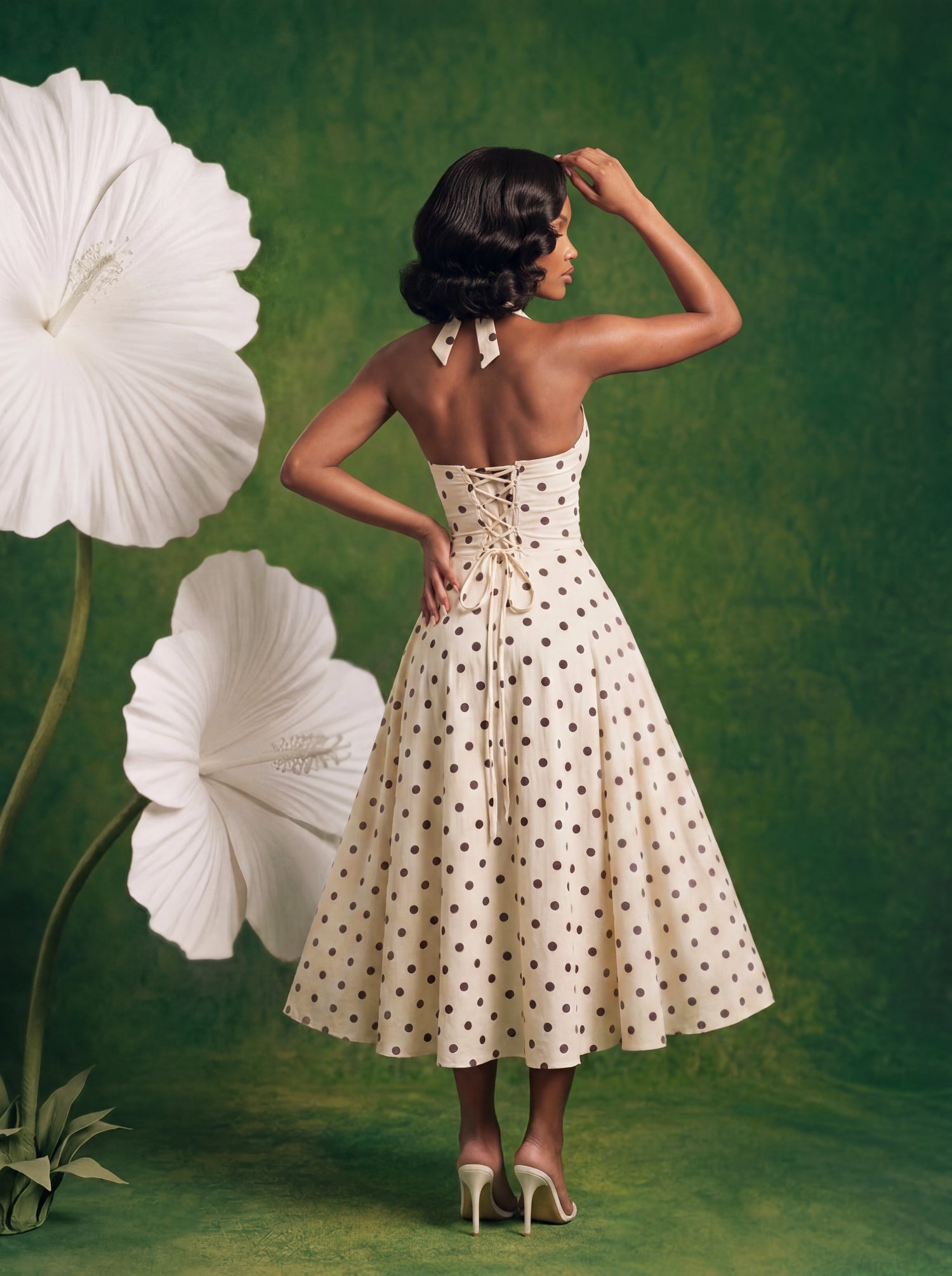 Back view of a cream polka dot halter midi dress featuring an adjustable lace-up corset back, flowing A-line skirt, and elegant open-back silhouette. Styled with oversized white flowers against a textured green backdrop.