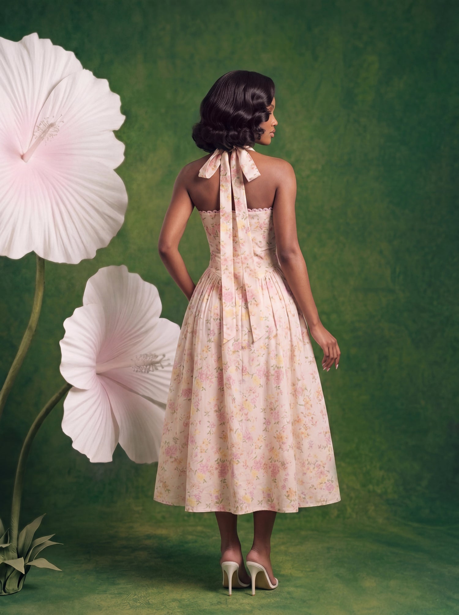 Back view of a soft pink floral halter midi dress featuring an elegant open-back silhouette with long self-tie halter straps and a flowing midi skirt. Styled with white heels against a textured green backdrop with oversized floral props.