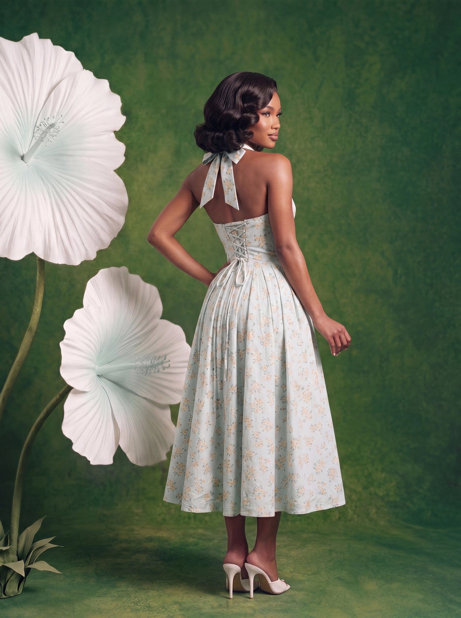 Back view of the light blue floral halter midi dress featuring an open back with adjustable halter ties and a lace-up corset closure. The full gathered midi skirt falls below the knees and is printed with a delicate yellow floral pattern throughout. The model is wearing white heels and standing against a green backdrop with oversized white flowers.