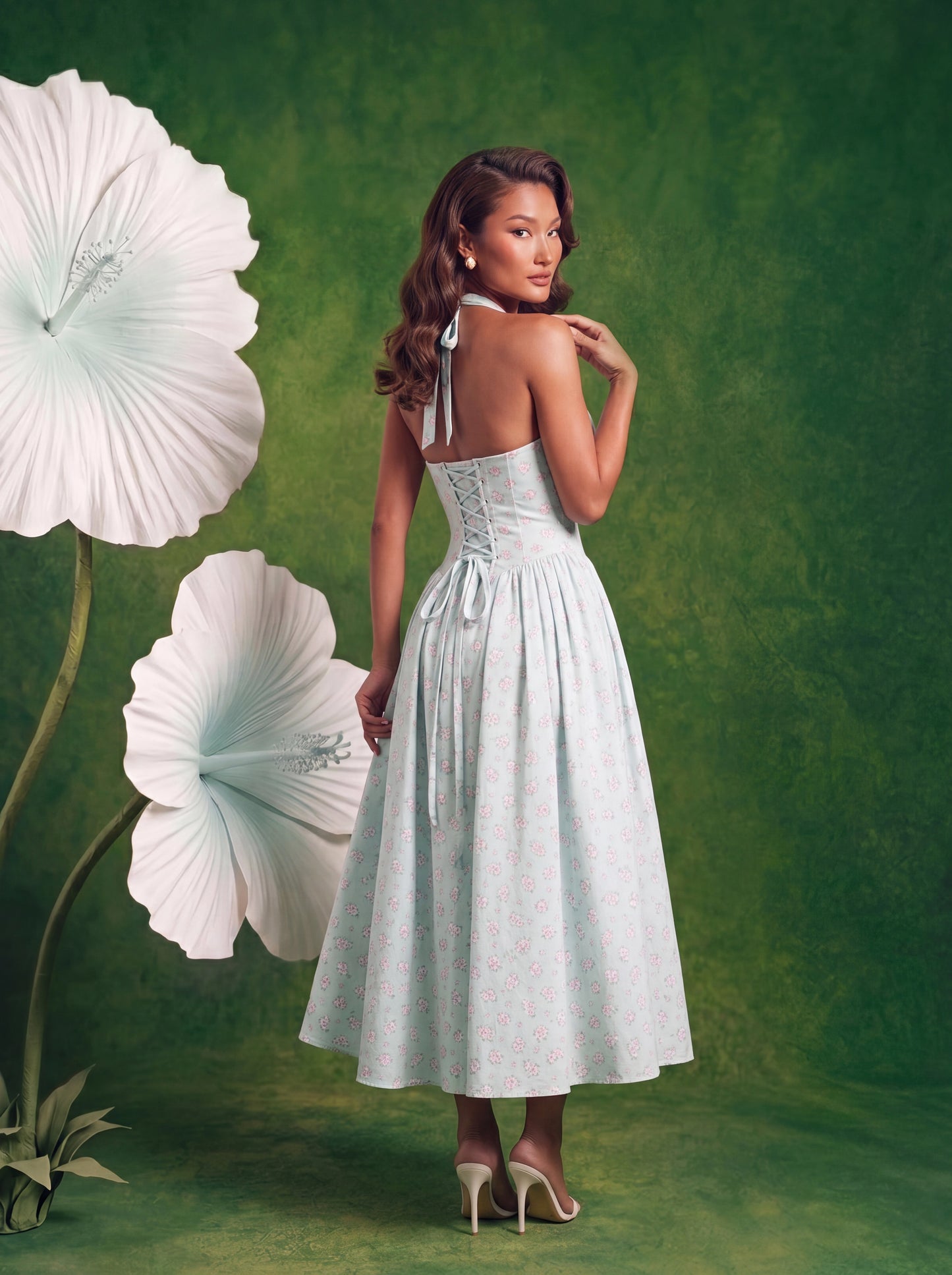 Back view of the light blue floral halter midi dress featuring an open back with adjustable halter ties and a lace-up corset closure. The full gathered midi skirt falls below the knees and is printed with a small pink floral pattern throughout. The model is wearing nude heels and standing against a green backdrop with oversized white flowers.