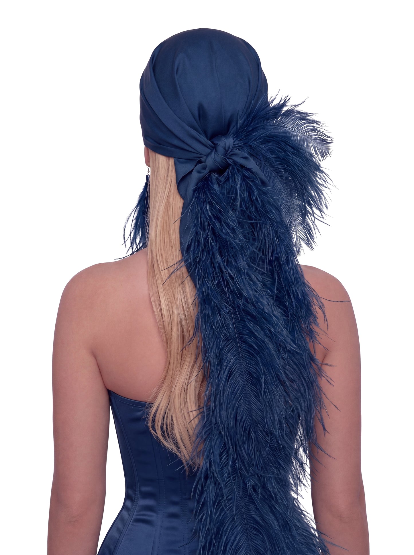A woman shown from the back wearing a navy blue satin headwrap or headband with a large feathered tail attached, styled over blonde hair against a plain white background.