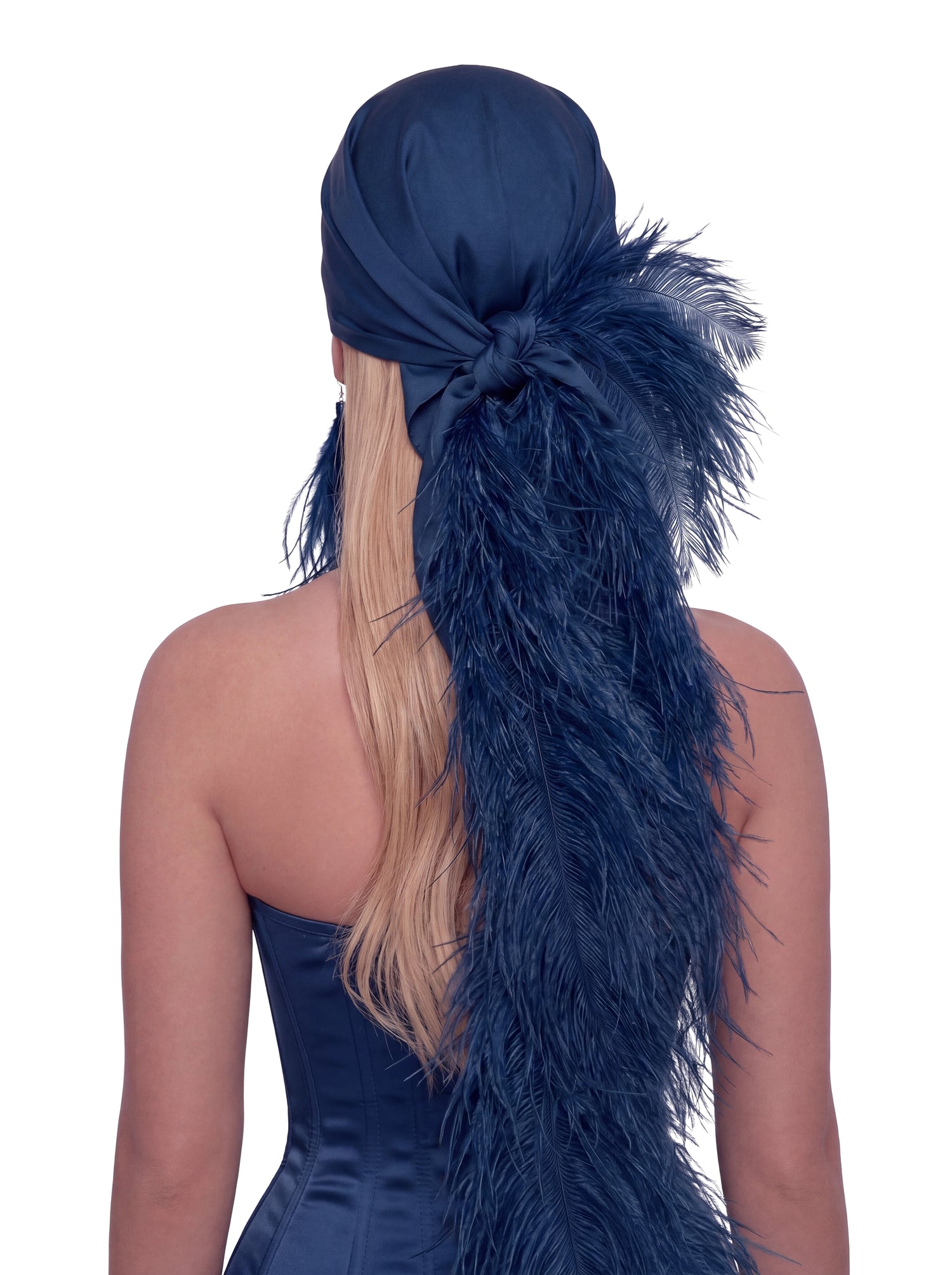 A woman shown from the back wearing a navy blue satin headwrap or headband with a large feathered tail attached, styled over blonde hair against a plain white background.