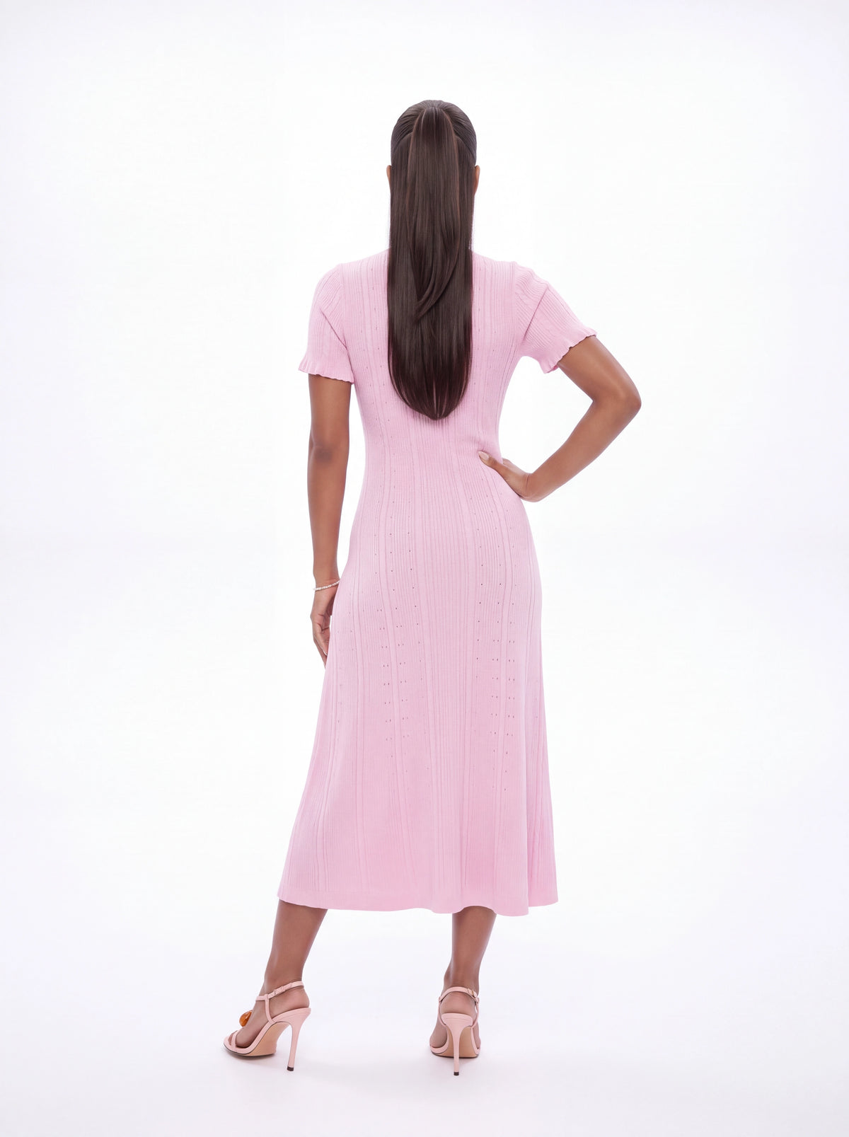 A woman viewed from behind wearing a light pink short-sleeve midi dress with a textured vertical rib pattern, paired with nude high-heeled sandals, against a solid white studio background.