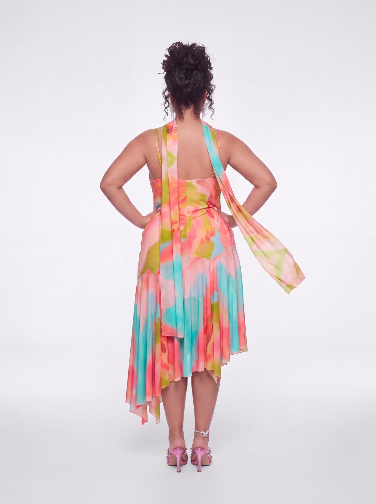 A woman shown from the back wearing a sleeveless halter dress with a colorful tie-dye pattern in pink, orange, yellow, and teal, paired with pink high-heeled sandals, against a light gray studio background.