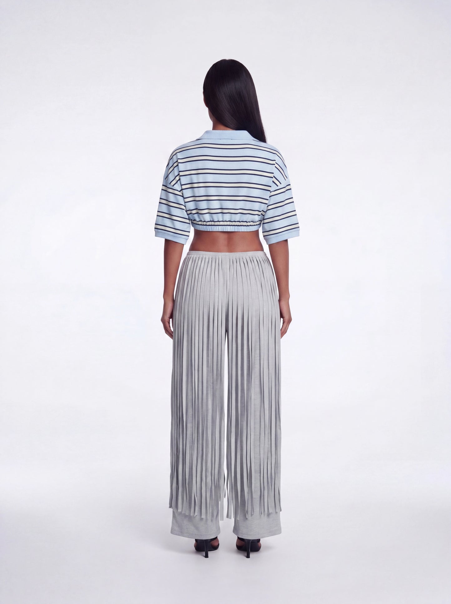 Back view of grey fringe wide-leg pants featuring layered fringe detailing and a stretchy waistband paired with a blue striped cropped collared knit top and black heels.