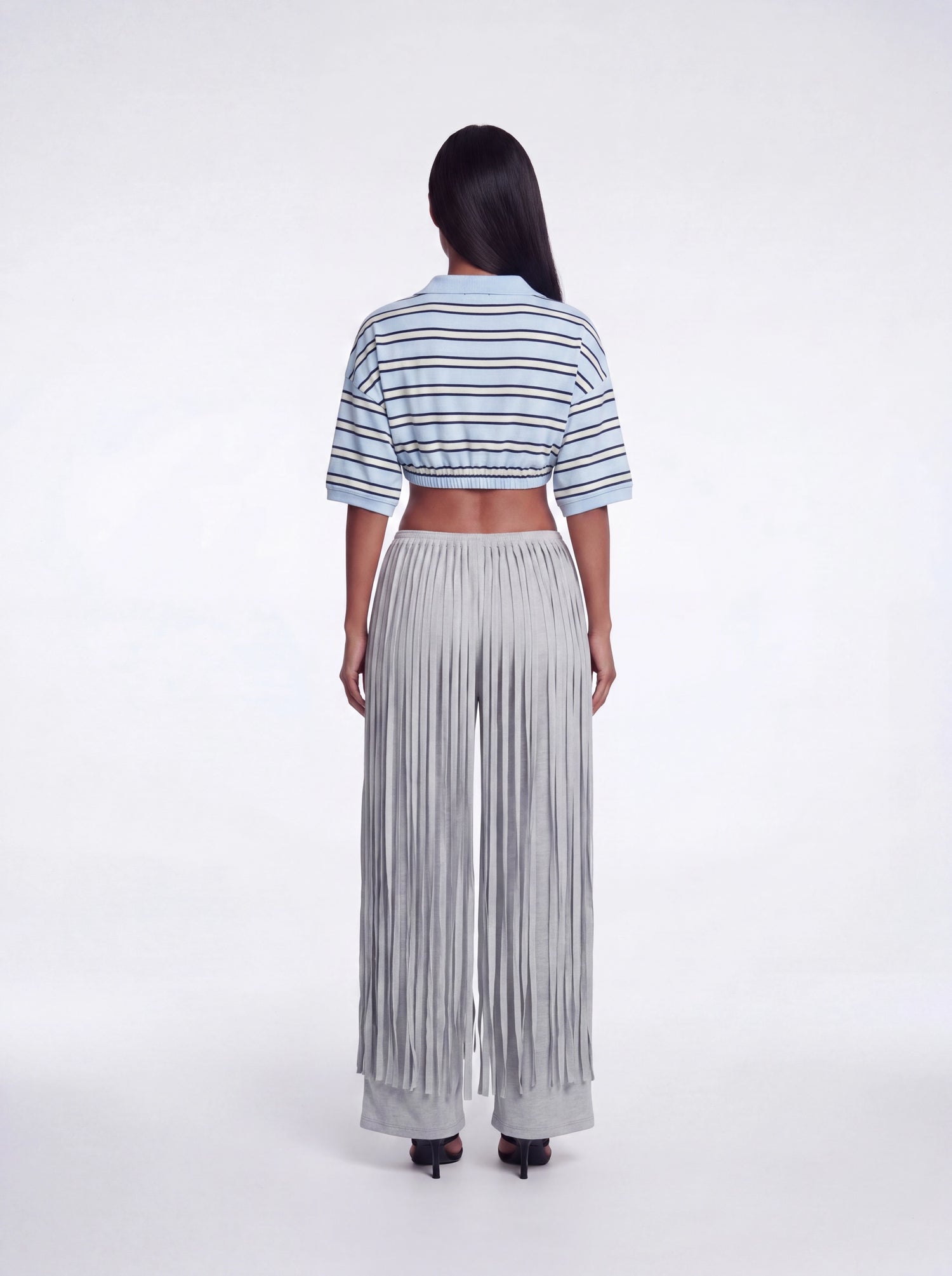 Back view of grey fringe wide-leg pants featuring layered fringe detailing and a stretchy waistband paired with a blue striped cropped collared knit top and black heels.