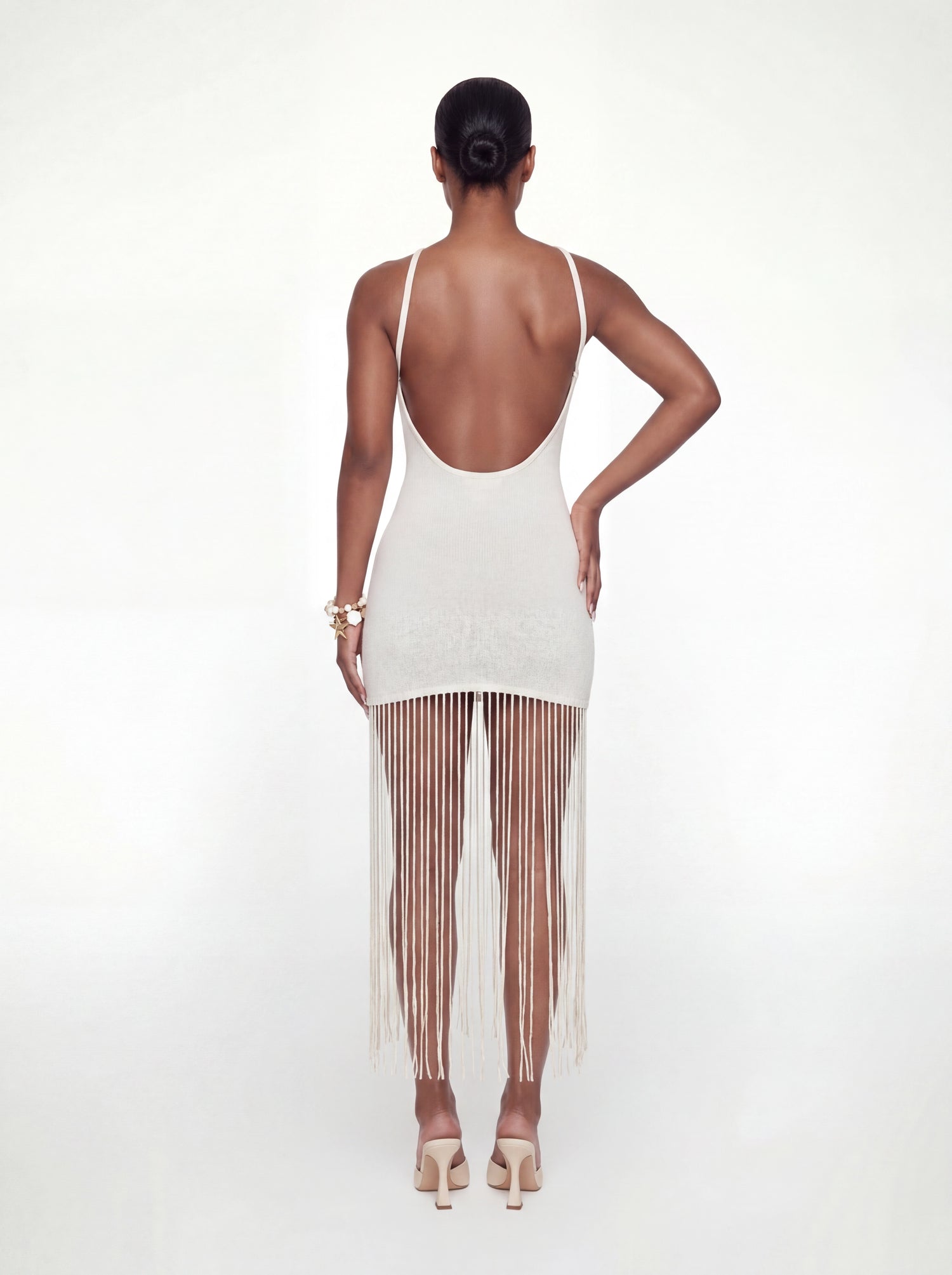 Back view of an ivory knit mini dress featuring a low open back, slim shoulder straps, a fitted silhouette, and a flowing long fringe hem. Styled with nude heeled sandals against a white backdrop.