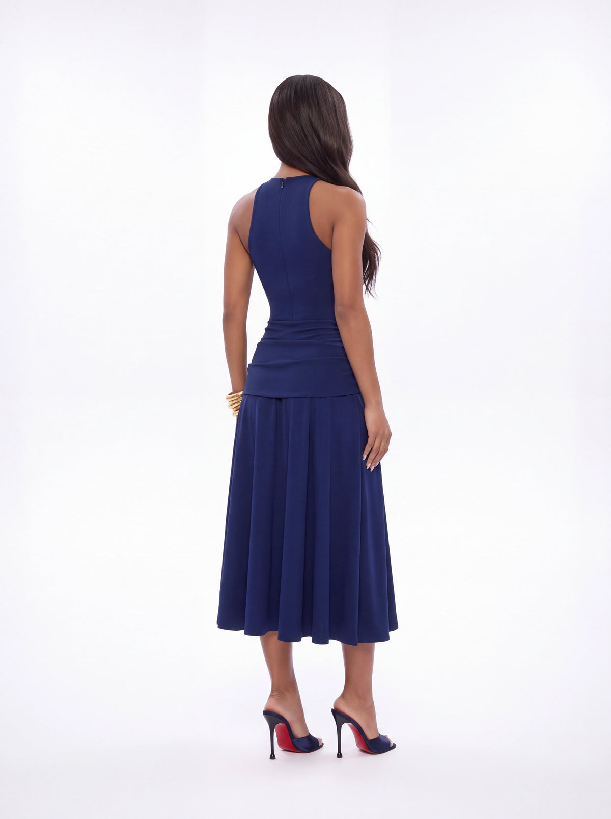 A woman viewed from the back wearing a sleeveless navy blue midi dress with a ruched waist and flared skirt, paired with navy heels, against a plain white studio background.