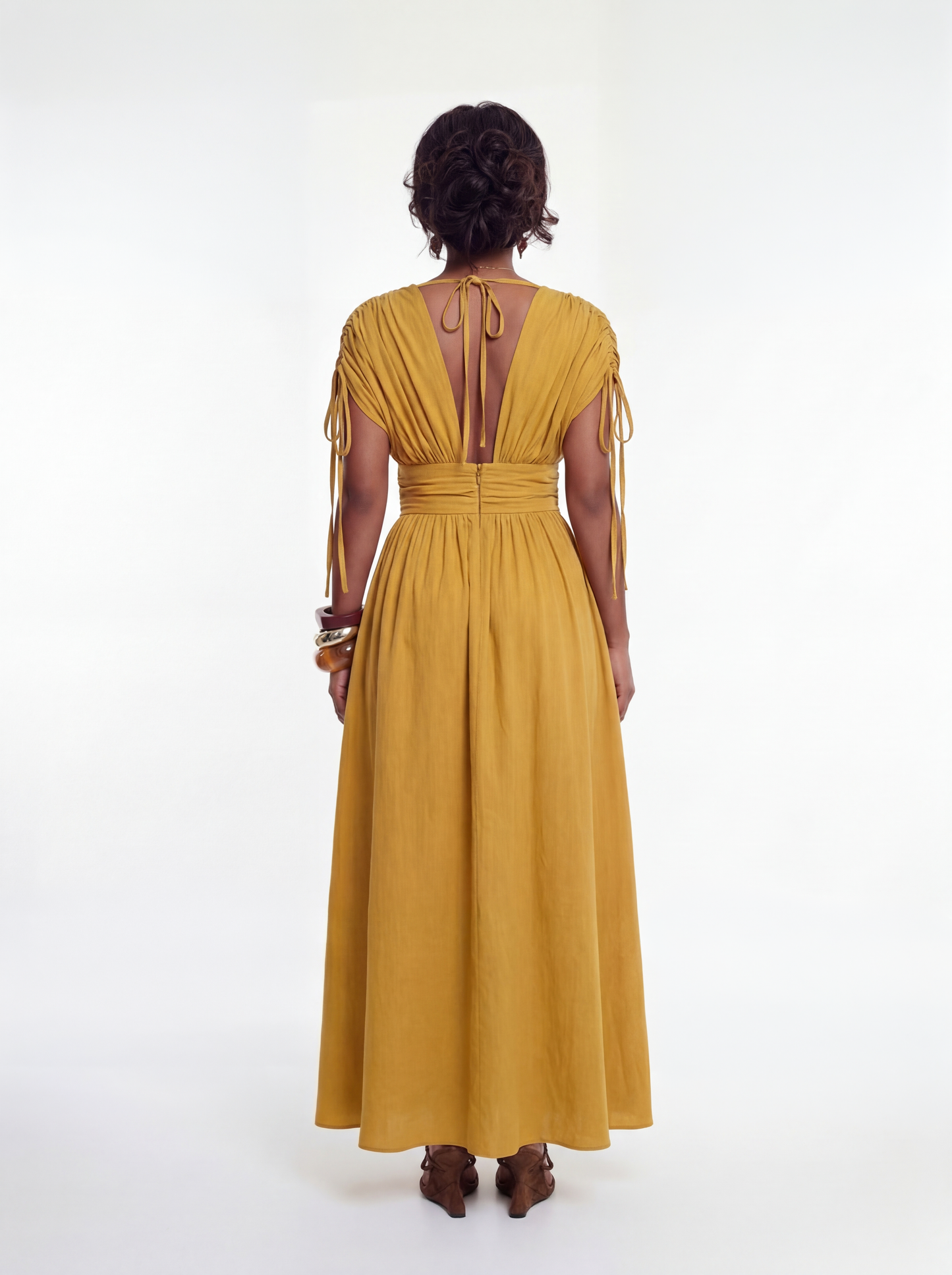Back view of a woman wearing a mustard yellow maxi dress featuring a deep open back with adjustable tie closure, ruched detailing along the bodice, a cinched waistband, and a flowing full-length skirt.