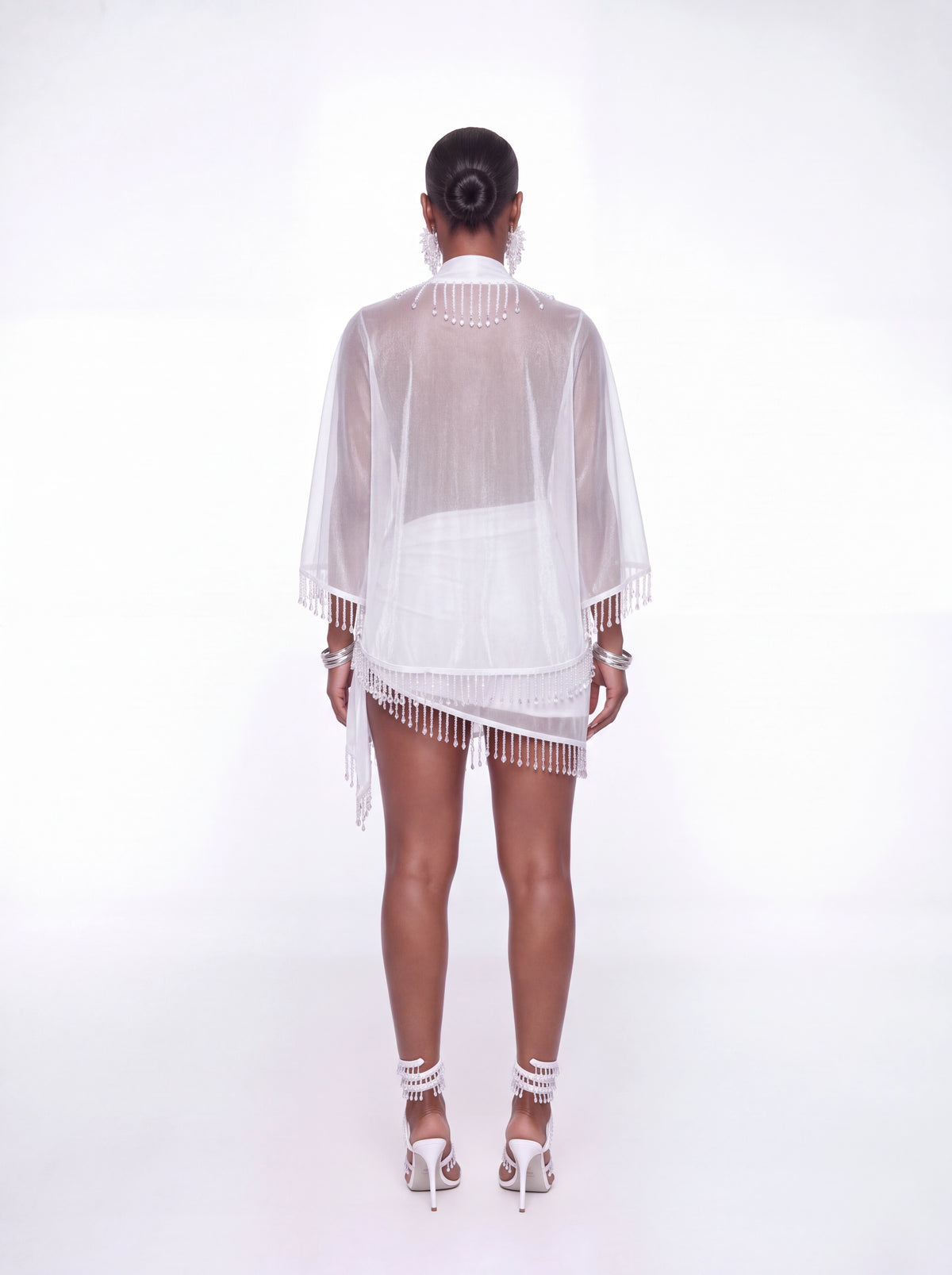 A model shown from the back wearing a sheer white fringed cover-up dress over a white mini dress, styled with white high-heeled sandals and silver jewelry against a white studio background.
