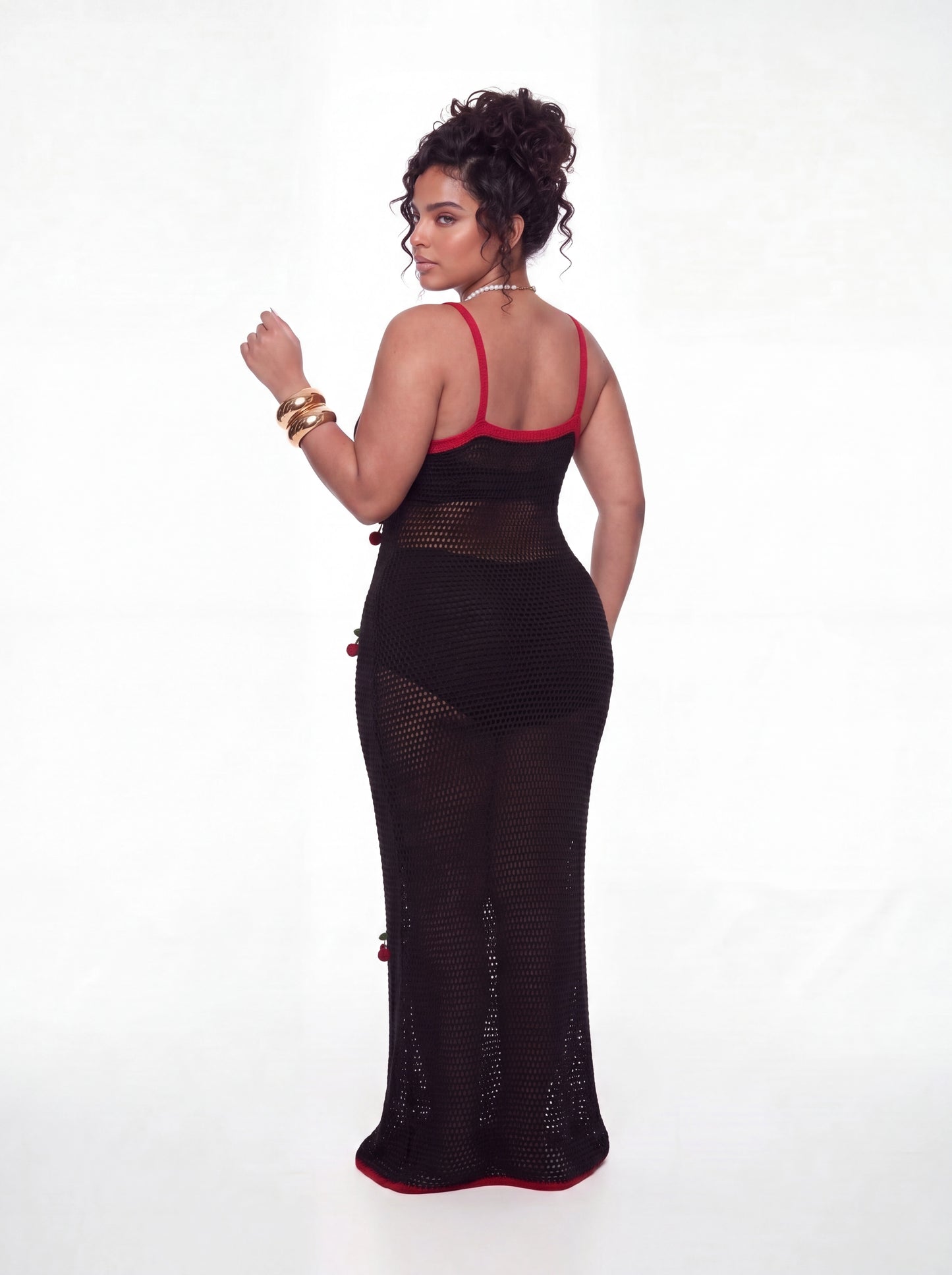 Curve model wearing a black open-knit crochet maxi cover-up dress featuring red contrast trim and three-dimensional cherry appliqués throughout. The fitted silhouette is layered over a black swimsuit and styled with gold bangles and a pearl cherry necklace against a white studio background.