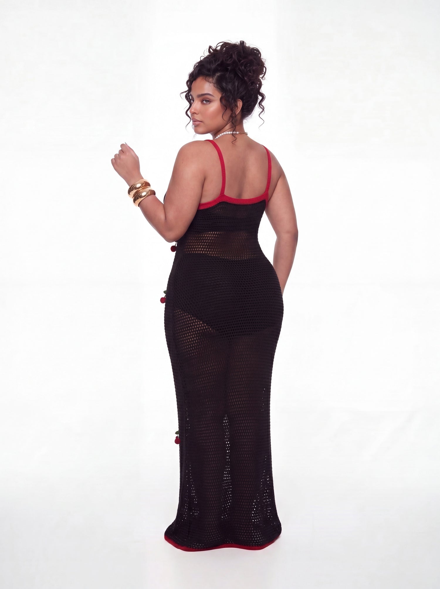 Curve model wearing a black open-knit crochet maxi cover-up dress featuring red contrast trim and three-dimensional cherry appliqués throughout. The fitted silhouette is layered over a black swimsuit and styled with gold bangles and a pearl cherry necklace against a white studio background.