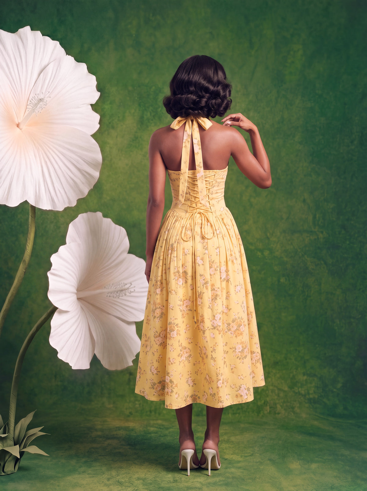 Rear view of a yellow floral-print midi dress featuring a lace-up corset back, adjustable halter neck tie, fitted waist, and flowing pleated skirt. Model poses in white heels against a green floral backdrop with oversized white hibiscus flowers.
