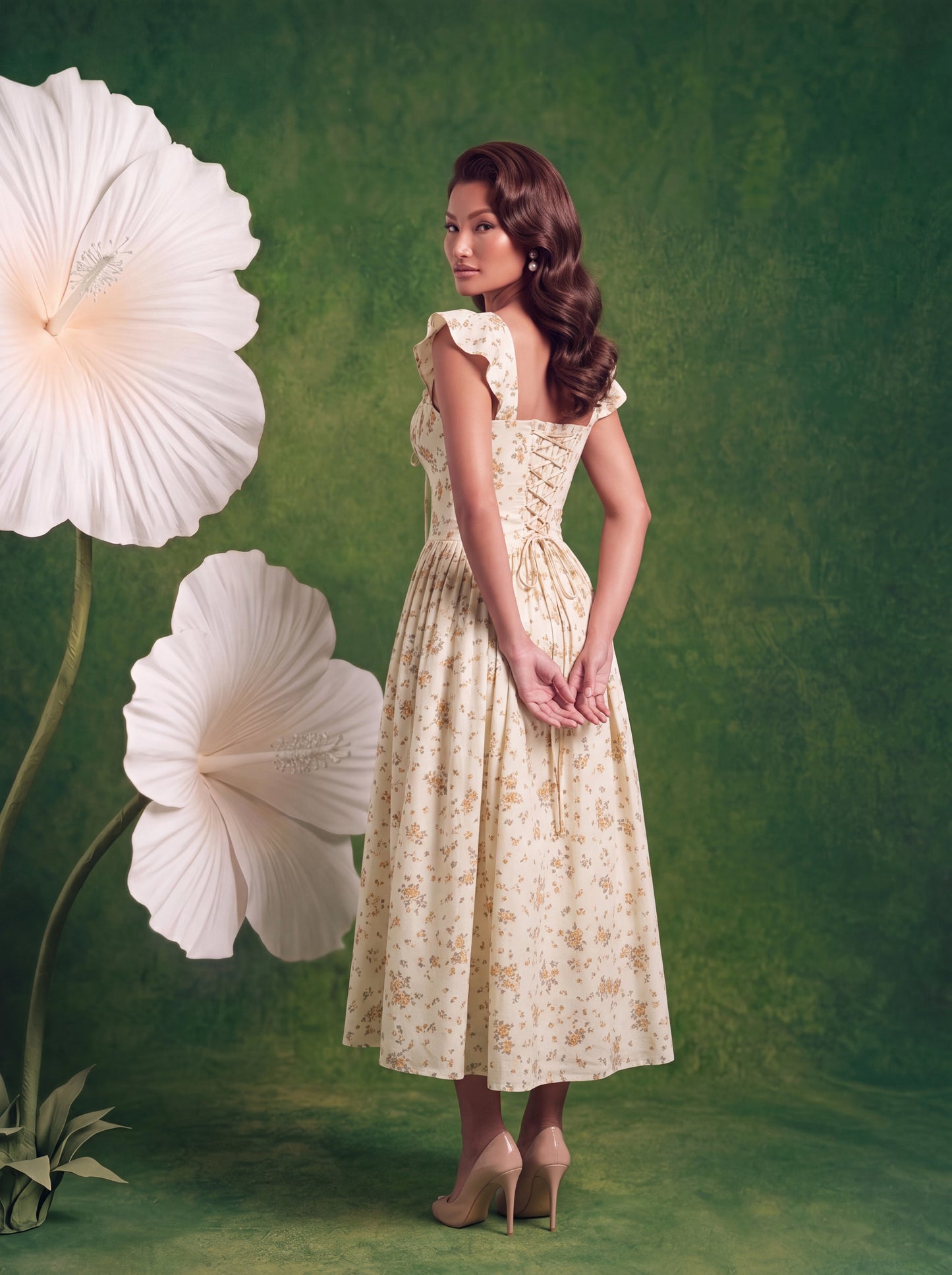 A woman poses from the back in a cream midi dress with a subtle yellow floral print, photographed against a green studio backdrop with oversized white hibiscus flowers. The dress features flutter sleeves, a lace-up corset back for an adjustable fit, and a flowing A-line skirt that falls below the knee. She completes the look with nude peep-toe heels, showcasing the elegant silhouette and feminine back detailing.