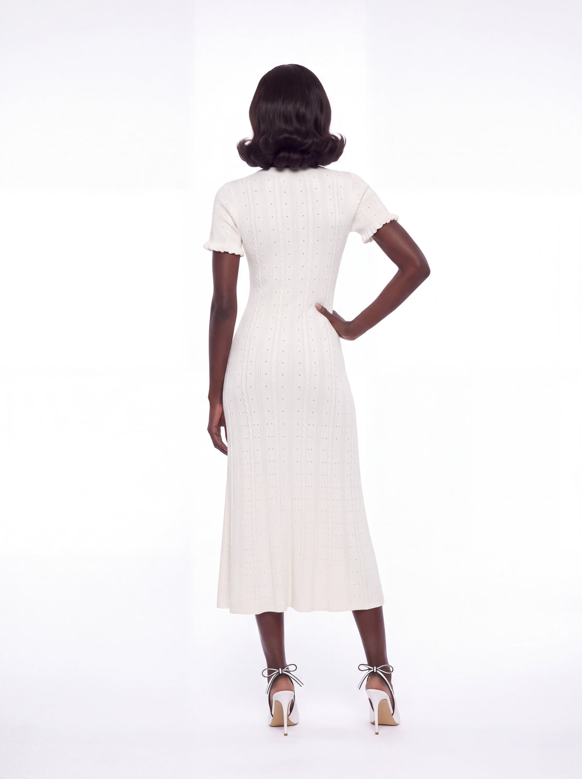 A model stands facing back against a seamless white studio background wearing a short-sleeved cream-colored textured midi dress with a button-down front and matching cream  heels with ribbon strings.