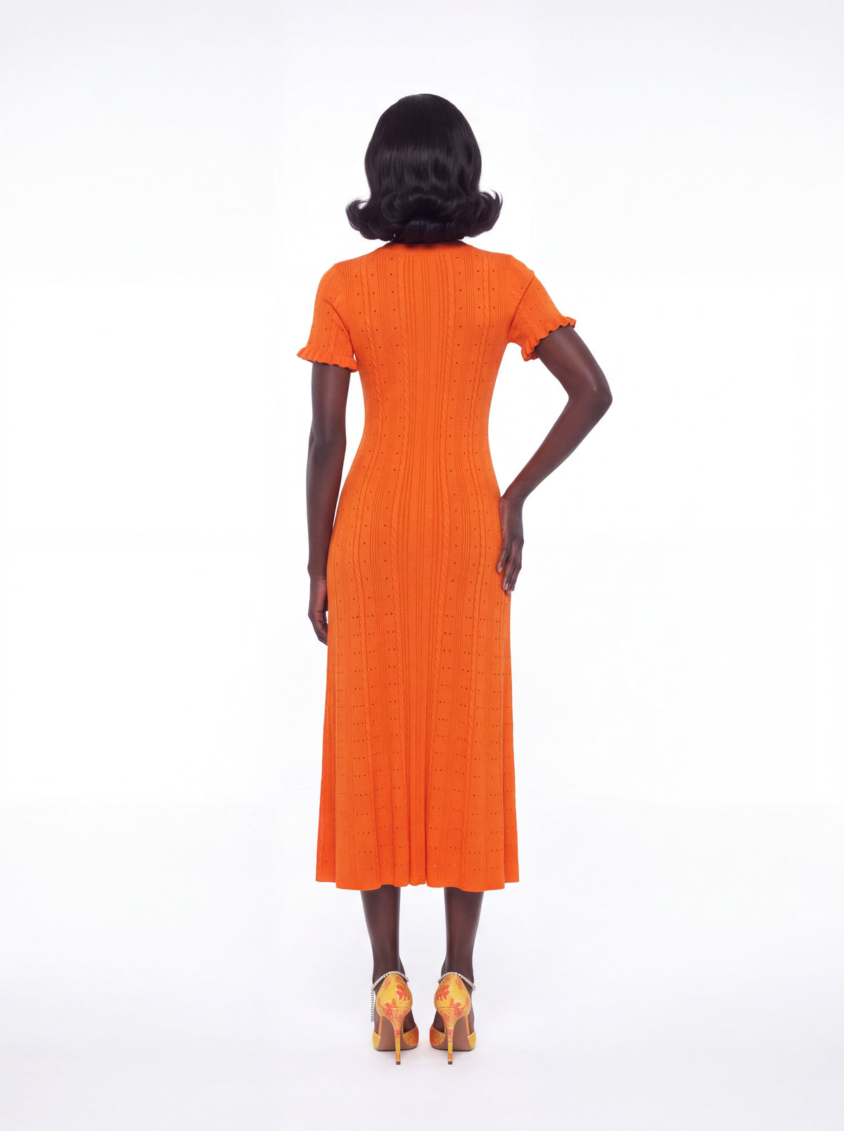 A woman viewed from the back wearing a bright orange short-sleeve midi dress with a textured vertical rib pattern and ruffled trim at the sleeves, paired with yellow high heels, against a plain white studio background.