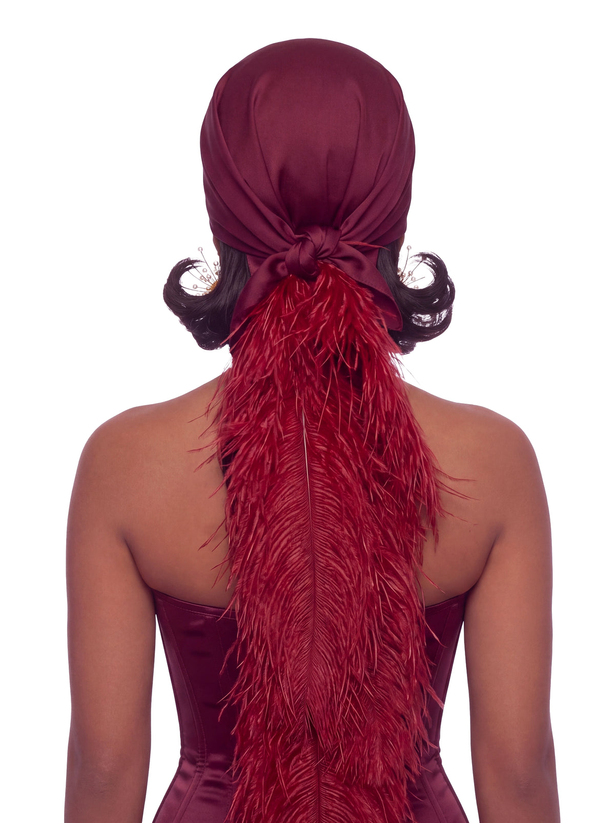 A model shown from the back wearing a deep burgundy satin headwrap or headscarf with a long matching feathered hair accessory, styled with curled dark hair and decorative hairpins against a plain white background.