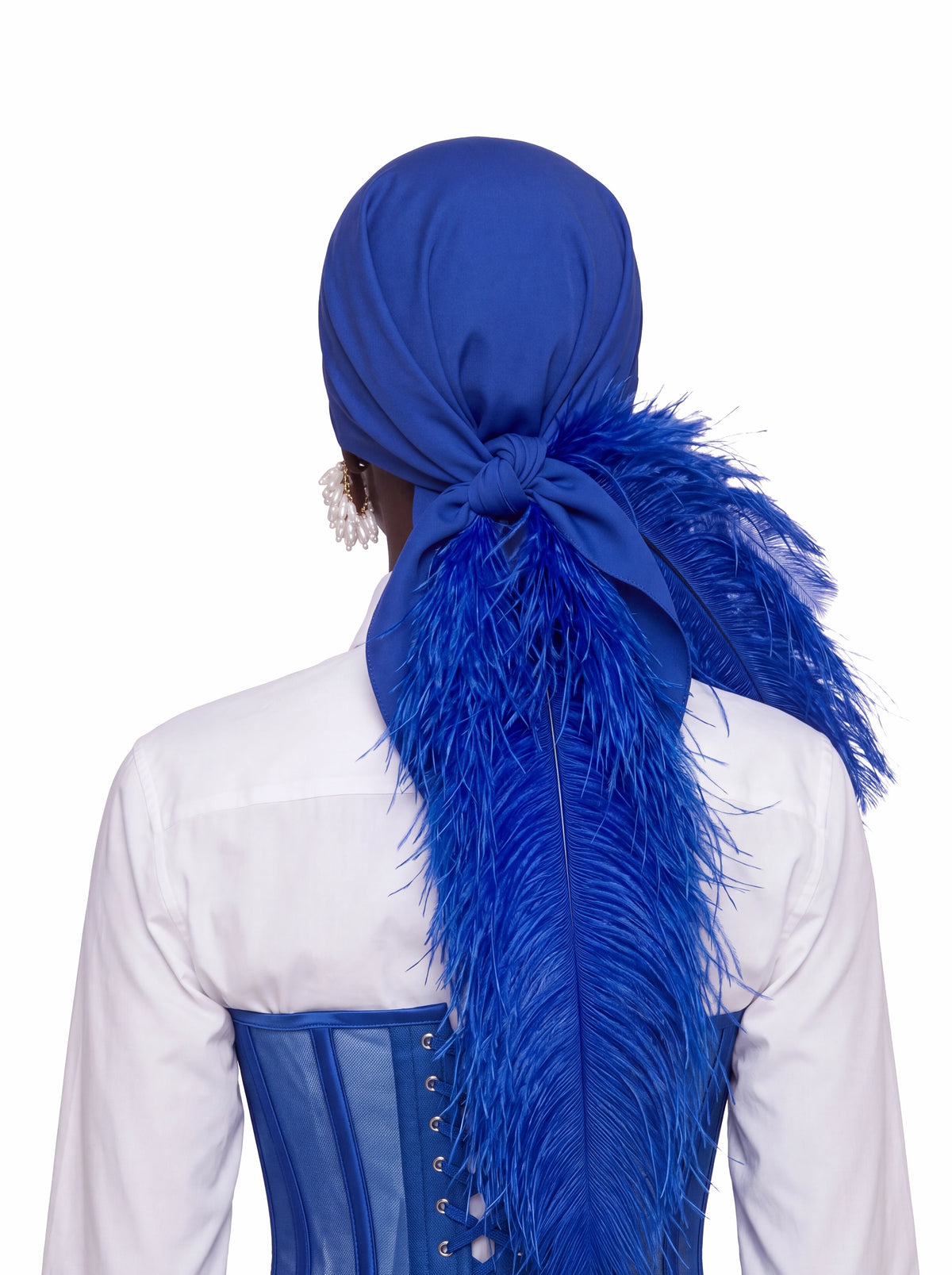 A person shown from the back wearing a royal blue fabric headwrap or durag with a large feathered accent hanging down the back, styled over a white shirt and blue corset against a plain white background.